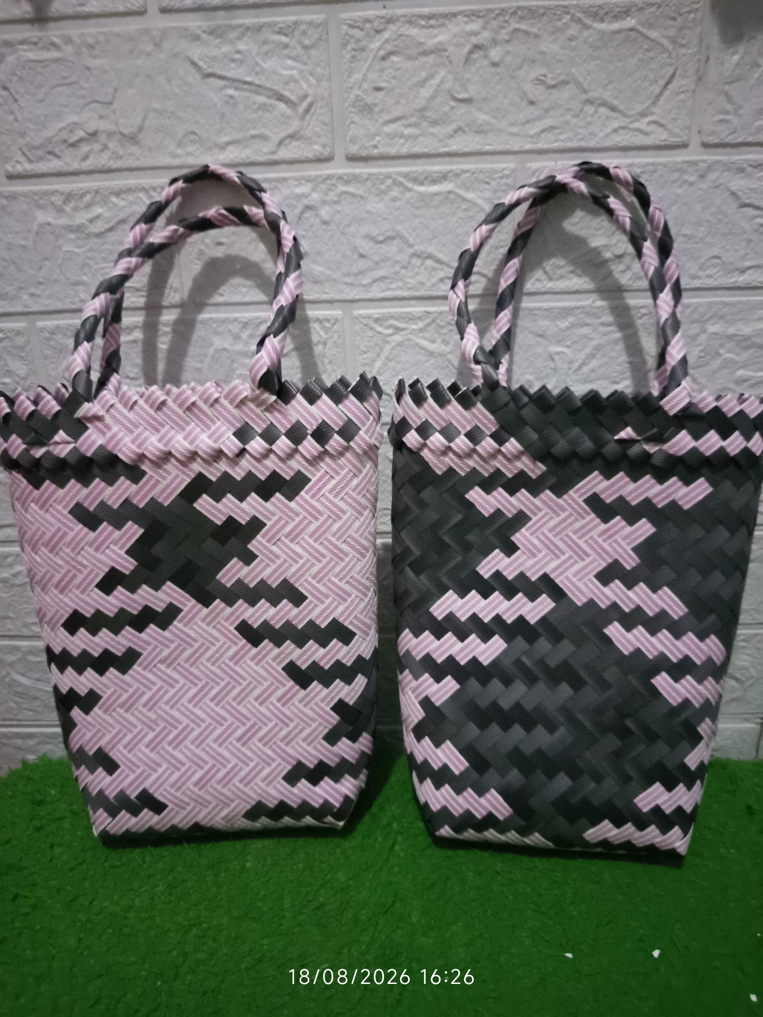 Two woven tote bags, one predominantly pink with dark grey accents and the other predominantly dark grey with pink accents, are displayed side-by-side against a white textured brick wall. Both bags feature braided handles and a geometric pattern woven into their bodies. They rest on a green, fuzzy surface. A timestamp, "18/08/2026 16:26," is visible in the lower right corner of the image. The scene appears to be a product display, possibly in a market or shop.