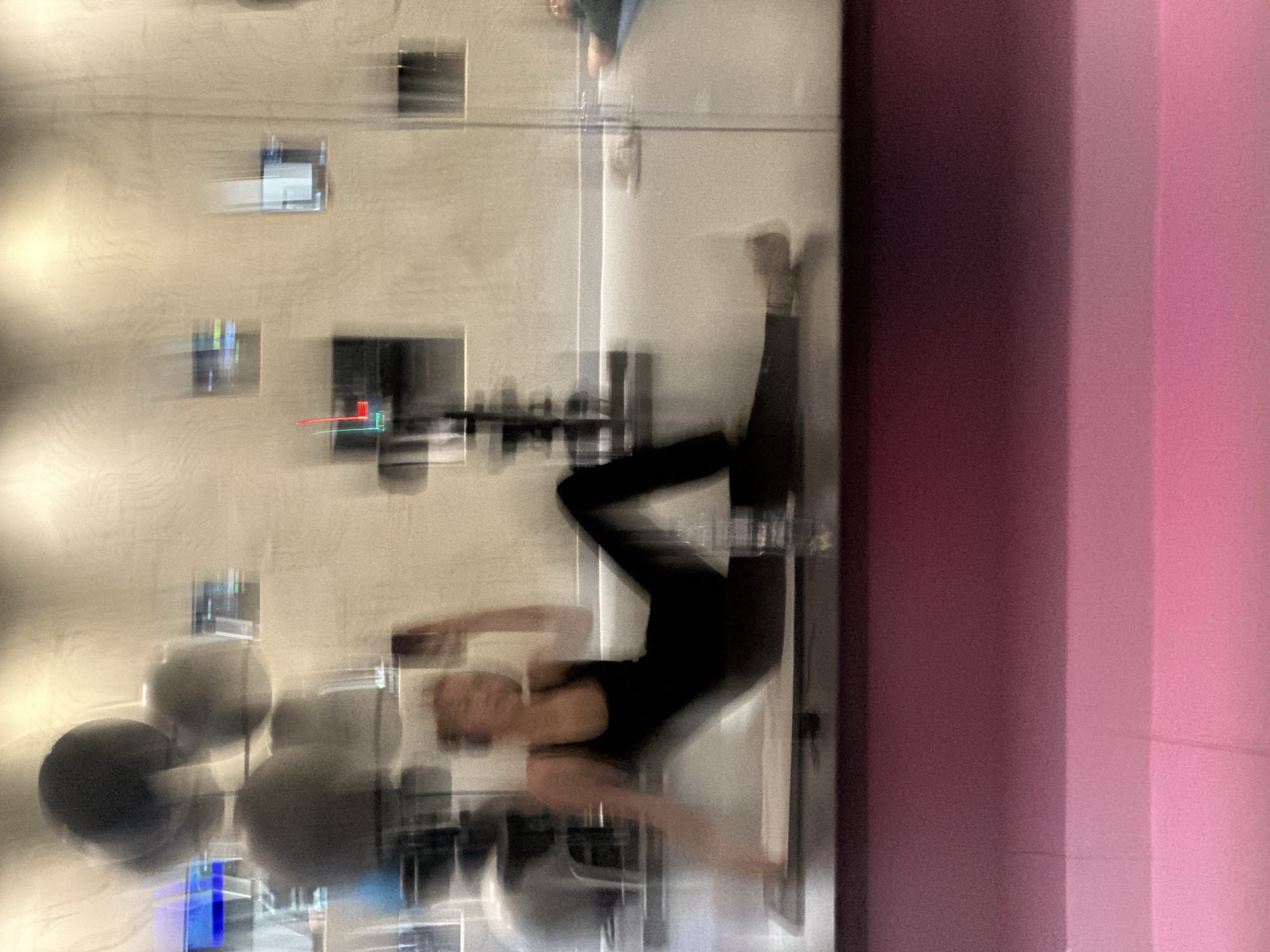 A blurry, slightly rotated image depicts a person in a gym. The person is wearing dark clothing and is in a contorted pose, possibly doing an exercise. The gym contains various exercise equipment, including medicine balls and weight racks. The walls are light colored with rectangular objects affixed to them. A multi-toned pink surface is visible on the right side of the image. The city and location are unknown.