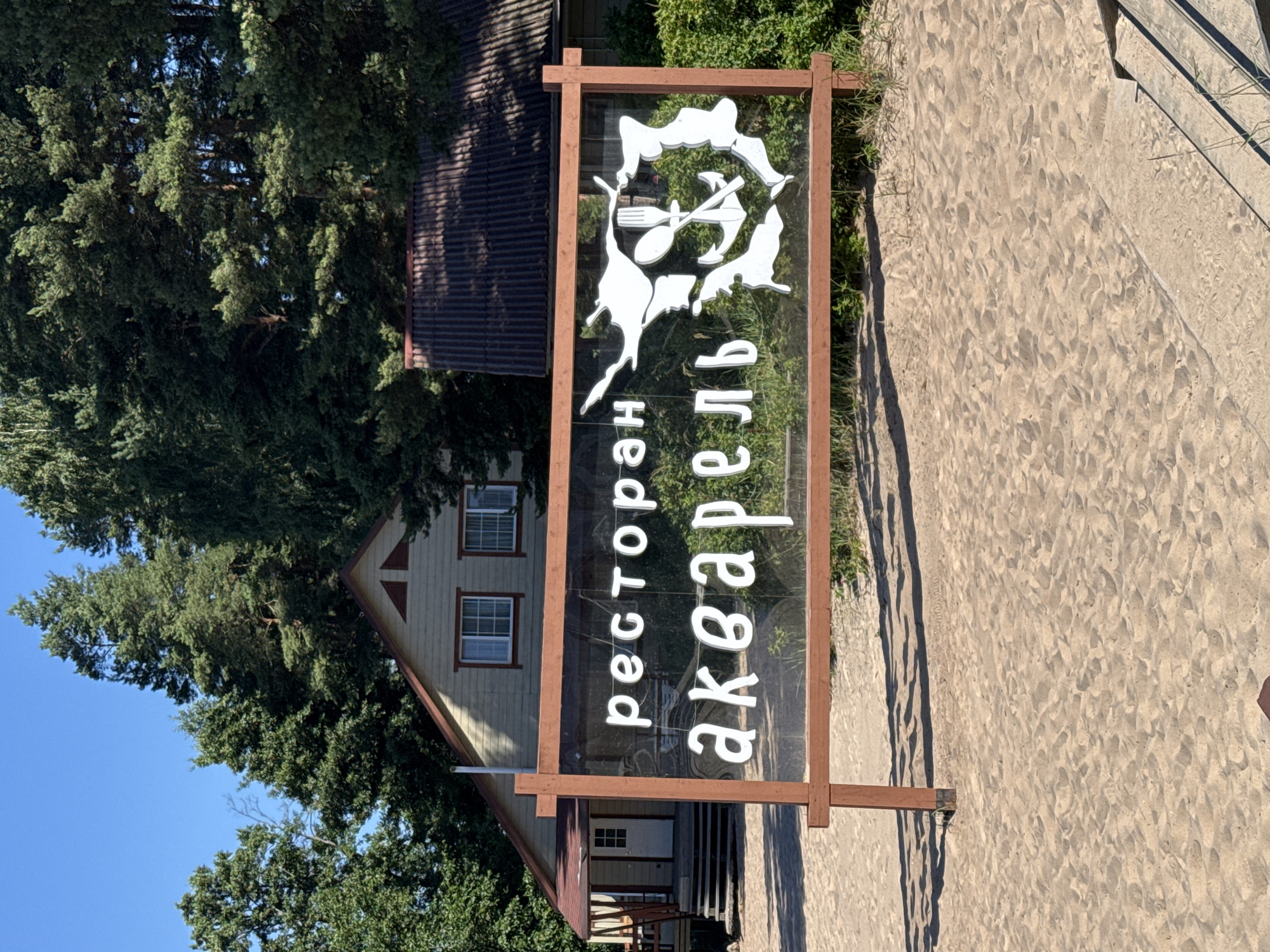 A wide outdoor shot captures a sunny day in Ozerki (Krasnodolinskaya), Russia, with a sandy foreground. A wooden-framed sign with glass is prominently displayed, featuring white Cyrillic text that reads "ресторан акварель" (restaurant watercolor). The sign also includes a white silhouette graphic of various nautical and culinary items, such as an anchor, fork, spoon, and possibly a fish. Behind the sign, a light-colored building with a brown roof and a porch is visible, surrounded by lush green trees and vegetation. The sky is a clear, bright blue.
