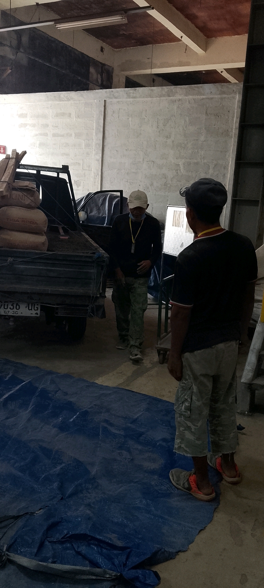Two men are in an indoor construction or warehouse environment in Depok, Indonesia. The man in the foreground, facing away from the camera, wears a dark t-shirt and camouflage shorts. He has a dark baseball cap on and is wearing slip-on shoes. The second man, in the middle ground, faces the first man and wears a dark long-sleeved shirt, camouflage pants, and a light-colored baseball cap with a yellow lanyard. He appears to be speaking or gesturing.
To the left of the men, a dark-colored flatbed truck is parked, loaded with several large sacks of what appears to be cement, possibly branded "Tiga Roda". Some wooden planks are also visible on the truck. A blue tarp is spread on the concrete floor in the foreground. The background consists of unfinished concrete block walls and a ceiling with exposed beams and lighting fixtures. The overall scene suggests an active work area or storage space.