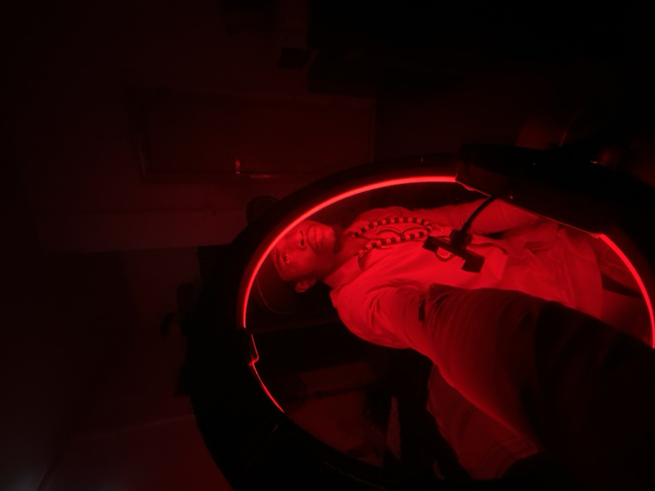Inside a dark room, a person lies within a circular device emitting red light. The subject is supine, wearing a light-colored shirt. The lighting casts deep shadows, obscuring details of the environment. A black apparatus is positioned above the person. The focus is on the individual within the illuminated ring, with the surrounding space largely undefined due to the limited light.