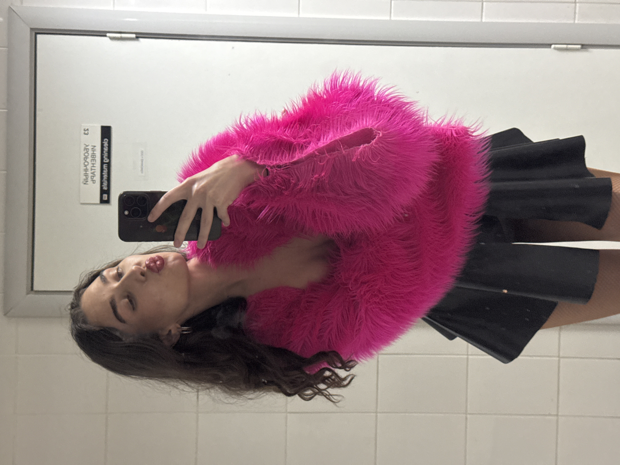 The image shows a young woman with dark, wavy hair taking a mirror selfie. She is wearing a bright pink, fluffy, long-sleeved top and a black mini skirt. Her lips are puckered as if for a kiss. In the background, the reflection shows a white tiled wall and a white door with two signs. One sign has Cyrillic text that appears to be backwards, possibly indicating it is a reflection of a sign on the other side of the door. The other sign is in English and reads "Theatrum Griselo." Based on the Cyrillic script, the location is likely Russia, and the reference to "Theatrum Griselo" could suggest a performance venue or theater, possibly within Kazan. The environment appears to be an indoor space, possibly a restroom or backstage area, given the tiled walls and utilitarian door.