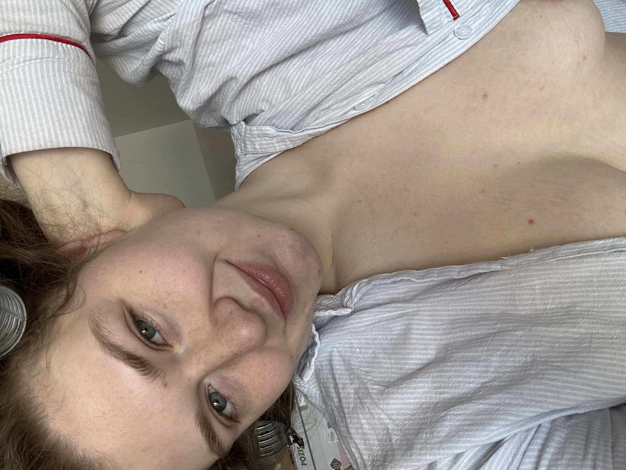A woman with fair skin, freckles, and light eyes is looking at the camera. She has light brown hair with metal hair rollers in it. She is wearing a light blue and white striped button-up shirt with red trim on the sleeve cuffs. Her head is resting on a white surface, possibly a bed or table. The image appears to have been taken indoors. There is no information available in the image to suggest this was taken in Kazan, Russia.