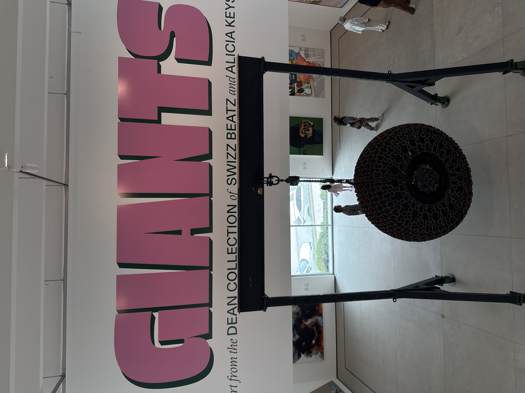 The image shows an art installation at the Museum of Contemporary Art San Diego, in San Diego, United States of America. The installation is suspended from a black metal crane structure and comprises a large, circular, chain-link form. The word "GIANTS" is displayed in large, pink, 3D letters, along with the text "Art from the DEAN COLLECTION of SWIZZ BEATZ and ALICIA KEYS". In the background, people can be seen walking through the gallery space, and paintings hang on the walls. Natural light streams in through large windows, illuminating the tiled floor.