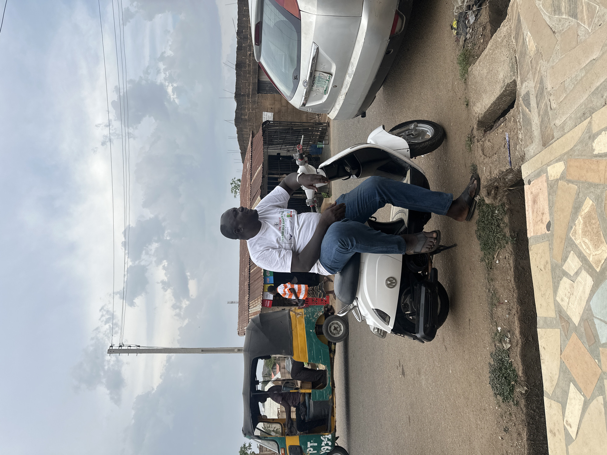 An outdoor daylight photo captures a street scene in what appears to be an urban or semi-urban area in Jos, Nigeria. The sky is overcast with clouds, and power lines run across the upper part of the frame.
In the foreground, a dark-skinned man is seated on a white scooter, facing left. He wears a white t-shirt, blue jeans, and dark flip-flops. His t-shirt has text that reads "THE VOICE OF THE YOUTHS FOR DEMOCRACY & GOOD GOVERNANCE" and "SUPER YOUTH MOVEMENT 2023" with a logo. He holds a small object in his right hand. The scooter has an "MM" logo on its front.
Behind the man and scooter, to the right, a white car (likely an SUV or hatchback) is parked, showing a Nigerian license plate that partially reads "JJ PLATEAU". The road is paved, and a patterned tiled sidewalk runs along the right edge of the frame, adjacent to a dirt patch and a concrete drainage culvert.
Further into the background, to the left of the scooter, a green and yellow tricycle, commonly known as a Keke Napep, is visible. A registration number, "PT-1934," is partially legible on its side. A person can be seen seated inside the tricycle. Beyond the vehicles, there are buildings constructed with brick walls and corrugated iron roofs, indicative of local architecture. Another person in an orange and white striped shirt is partially visible near one of these structures. A concrete utility pole supports the power lines that crisscross the sky.