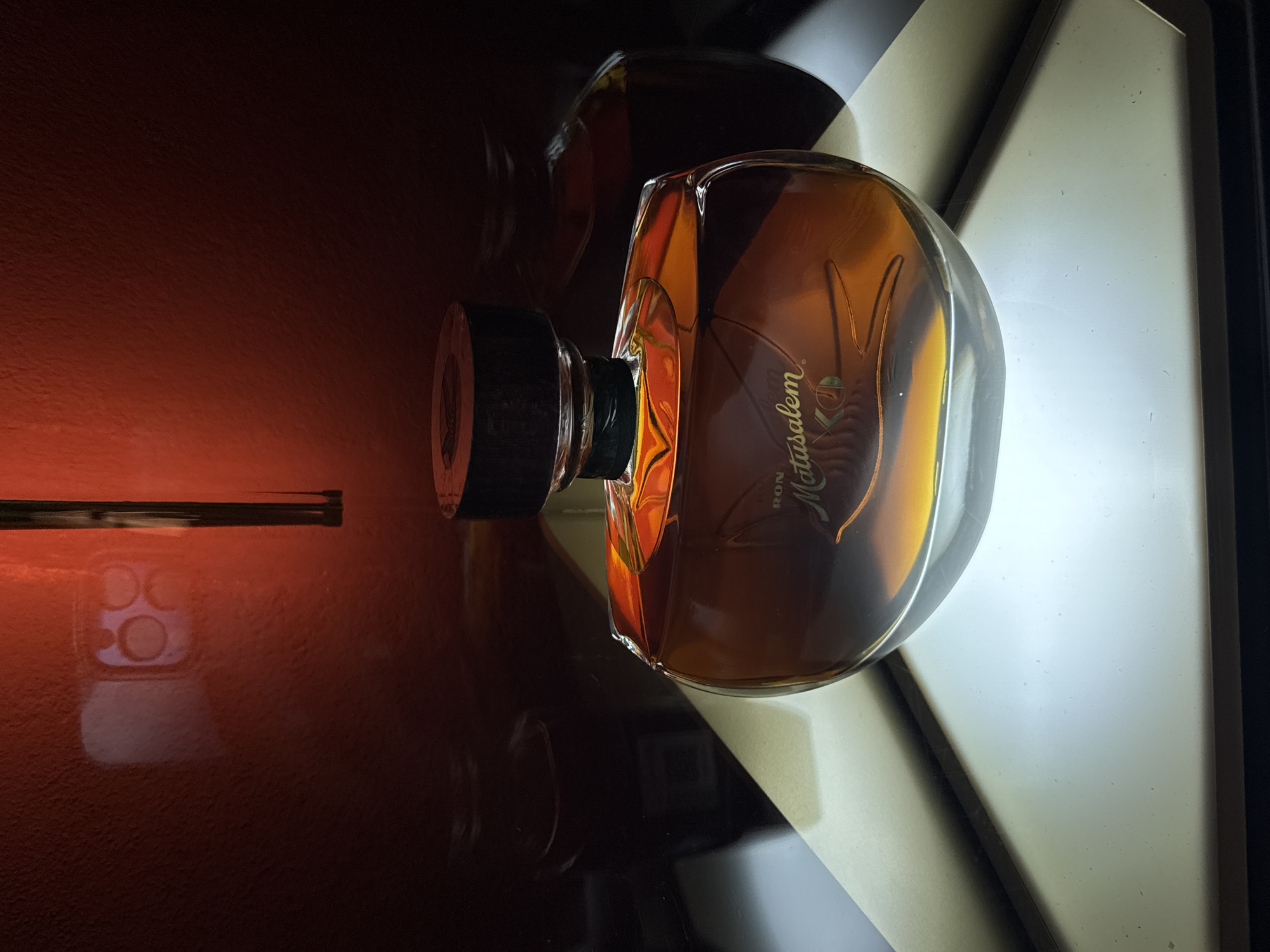 The image depicts a bottle of Ron Matusalem rum positioned on a glossy surface, likely a table. The bottle is oriented horizontally, with the cap towards the left and the main body towards the right. The background is split into two distinct sections: a dark, reddish-brown area on the left, reflecting the bottle, and a bright, white area on the right, illuminating the bottle. The light creates a sharp contrast and highlights the bottle's shape and contents. In the reflection on the left, there's a hint of a smartphone camera. There's nothing to indicate this image was taken in San Bernardino Tlaxcalancingo, Mexico.