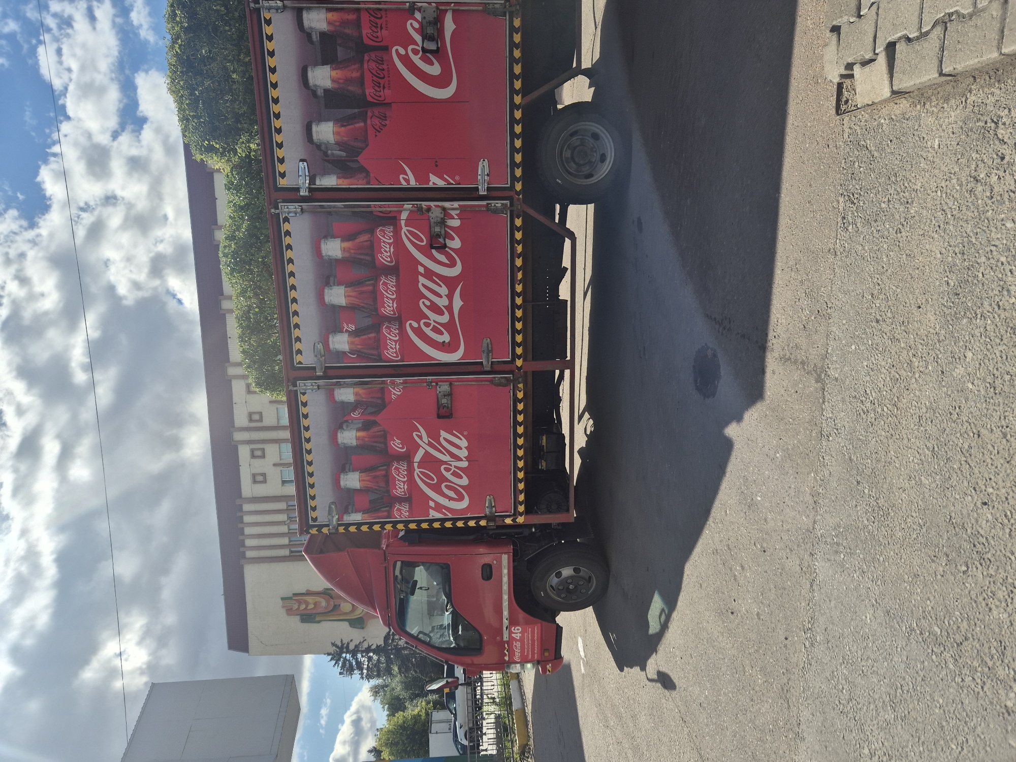 A red Coca-Cola delivery truck is parked on a paved surface, likely a street or parking lot, in Kokshetau, Kazakhstan. The truck's cargo area is open, revealing multiple cases of Coca-Cola bottles. The side of the truck prominently displays the Coca-Cola logo in white lettering against a red background. The truck has "Coca-Cola 46" written on the driver's side door. In the background, a building with a distinct architectural style, featuring vertical beige columns and square window-like elements, is visible. Trees and a partially visible white structure are also in the background. The sky is partly cloudy. The scene appears to be captured during daylight hours.