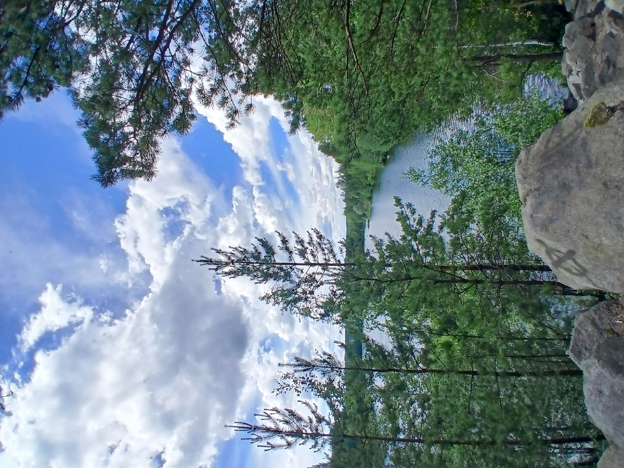 The image depicts a serene natural landscape featuring a body of water bordered by lush green trees. The sky above is a vibrant blue, adorned with dramatic white clouds. In the foreground, large gray boulders punctuate the scene, hinting at a rocky terrain. The overall impression is one of tranquility and unspoiled nature, likely a park or natural reserve. Without further information or context, it is impossible to determine the specific location within Yekaterinburg, Russia.