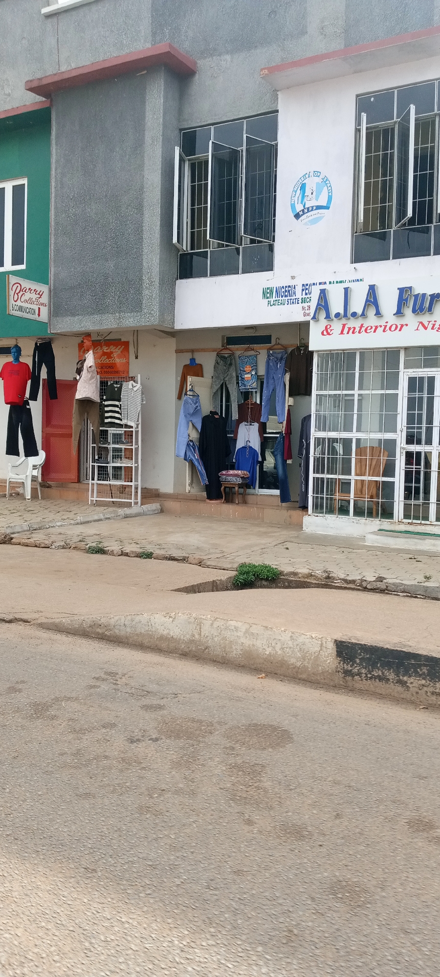 This image presents a street-level view of a commercial building in Jos, Nigeria, under bright daytime conditions. The building's facade is segmented, featuring a green section on the left, a grey middle portion, and a white section on the right, all with windows, some of which are open and fitted with metal grilles.
On the left, a shop identified by a red sign as "Barry Collections & Communication" displays clothing on two mannequins; one blue figure wears a red t-shirt and black pants, while an orange figure displays a light-colored shirt and khaki pants. A partially visible sign below the main one reads "Barry Collections" along with a phone number beginning with "0803". A white plastic chair sits on the sidewalk nearby.
The central and right sections of the building feature a political party presence. A prominent blue and white logo of the "NEW NIGERIA PEOPLES PARTY NNPP" is affixed to the white wall, accompanied by the slogan "Front Square and Proper." Below this, text reads "NEW NIGERIA PEO / PLATEAU STATE SECR / No. 28 / Gow," indicating a state secretariat office.
Adjacent to this, another shop, "A.I.A Furni & Interior Nig," is visible, with its name in blue lettering above a glass entrance. Outside the shops, various garments, including jeans, shirts, and dresses, are hung on racks and hangers, suggesting additional clothing businesses. A small upholstered stool is also seen on the pavement.
The foreground shows a paved asphalt road and a concrete sidewalk with distinct curbs, where small patches of green vegetation grow. The clear, even lighting and lack of strong shadows indicate it is likely midday or early afternoon with pleasant weather. The scene depicts a typical commercial street with a mix of retail and political offices.