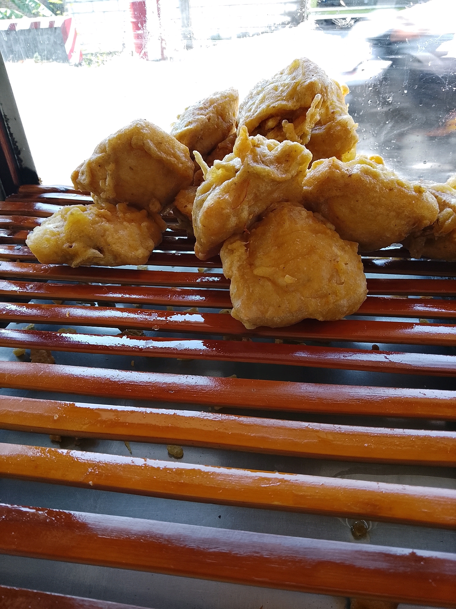 A close-up, eye-level shot shows a pile of fried snacks, possibly pisang goreng (banana fritters), arranged on a slatted wooden surface. The fritters are golden brown and appear to be coated in a batter, with some showing irregular shapes and textures. The wooden slats are dark reddish-brown and have a glossy finish, with some appear to be wet or sticky. Behind the fritters, a blurred background suggests an outdoor environment, possibly a street scene in Banaran, Indonesia, with sunlight filtering through a window or glass. The lighting is bright, creating highlights on the fritters and the wooden surface. The overall impression is of a food stall or street vendor display.