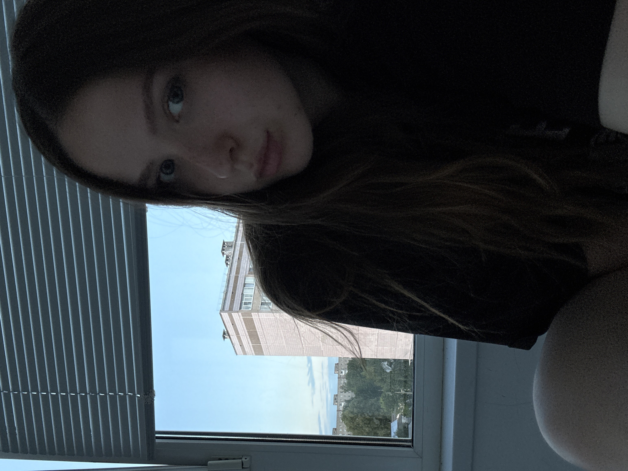 A young woman with long brown hair is in the foreground, looking towards the camera. She is wearing a dark top. Behind her, through a window covered by horizontal blinds, a pale blue sky is visible, with some clouds. In the background, there is a tall, multi-story building with a light-colored facade, and further in the distance, other buildings and trees can be seen, suggesting an urban or suburban environment. The image appears to be taken indoors, likely in an apartment in Chaikovsky, Russia, given the architectural style of the buildings.