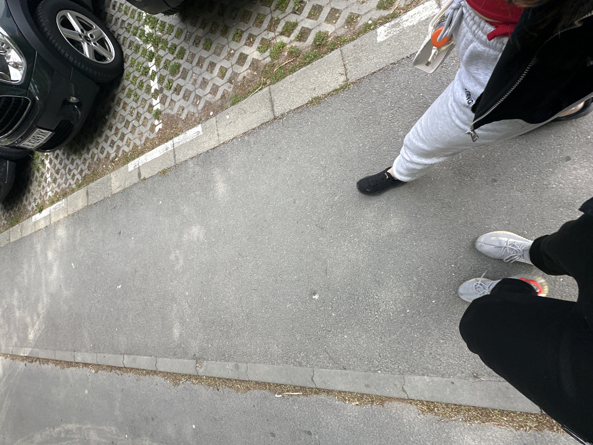 The scene shows an outdoor street in Varna, Bulgaria, adjacent to a parking area paved with grasscrete. A dark green car partially occupies the left side of the frame. Two individuals are present, both visible from the waist down. One person wears gray sweatpants, a red top, a black jacket, and black sneakers. The other person wears black sweatpants and gray Yeezy sneakers with a hint of orange on the sole. The individuals are standing on the gray asphalt of the street. The street is bordered by a concrete curb with some debris and vegetation along the edge.