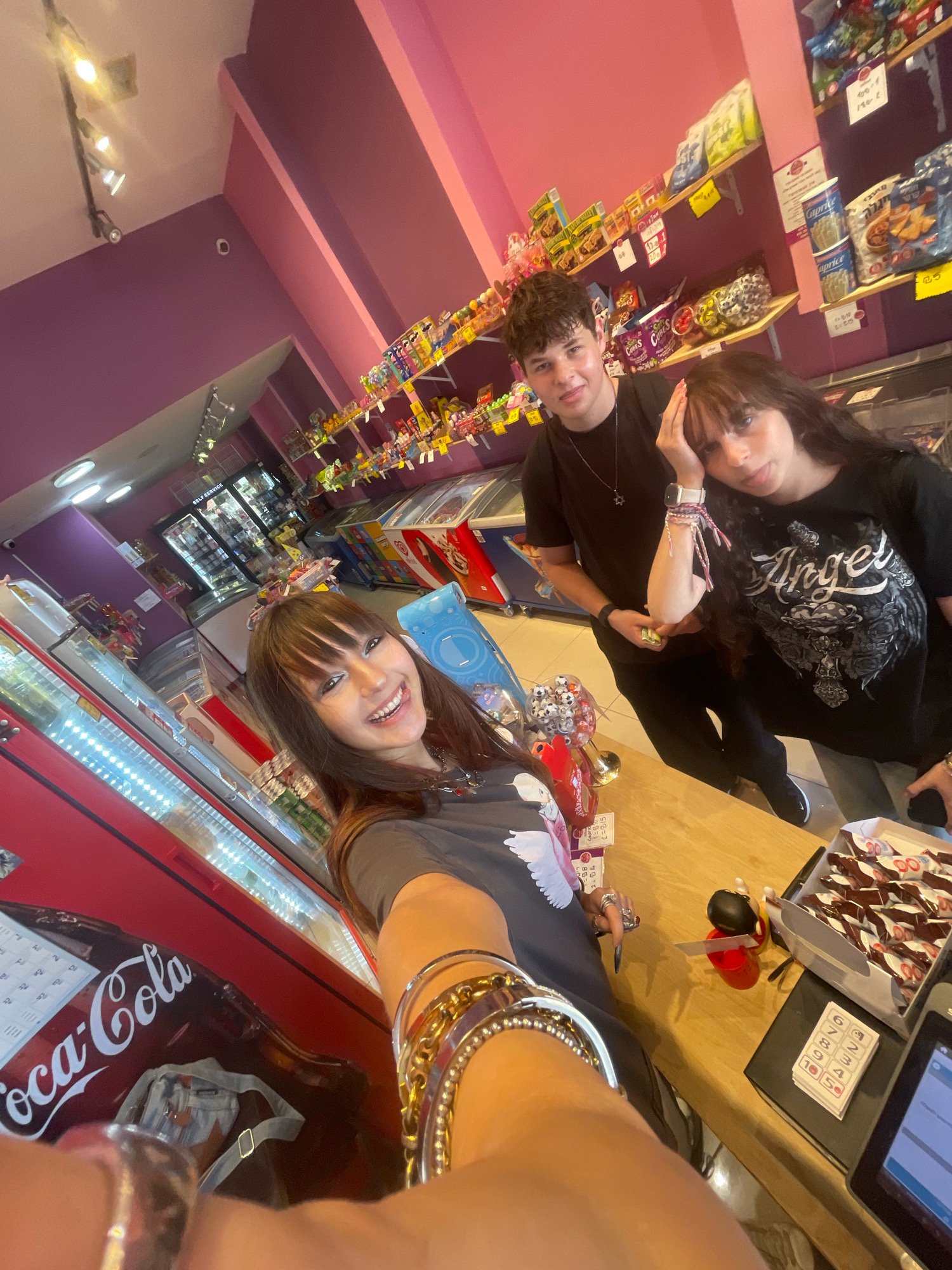 Three individuals, two young women and one young man, are posing for a selfie in a brightly lit convenience store. The store features shelves stocked with snacks and candies, and refrigerated display cases. The walls are painted in shades of pink and purple, and the lighting is provided by track lights on the ceiling. The individuals are dressed casually. One woman is in the foreground, holding up her arm, and smiling at the camera. The other two are standing behind her. The overall scene suggests a casual, everyday outing.