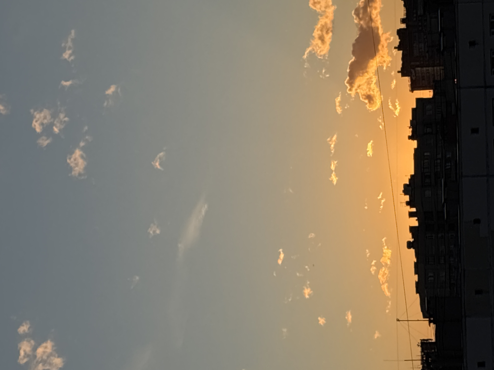 The image shows a silhouette of apartment buildings against a sunset sky. The sky transitions from a pale blue at the top to a warm orange and yellow near the horizon, indicating dusk. Scattered clouds are visible, some illuminated by the setting sun. The buildings are dark and their architectural details are not clearly discernible due to the low light. They appear to be multi-story residential structures typical of an urban environment. The scene is devoid of any people or visible actions. The overall impression is a quiet evening in a city, likely in Russia, given the architectural style suggested by the buildings.