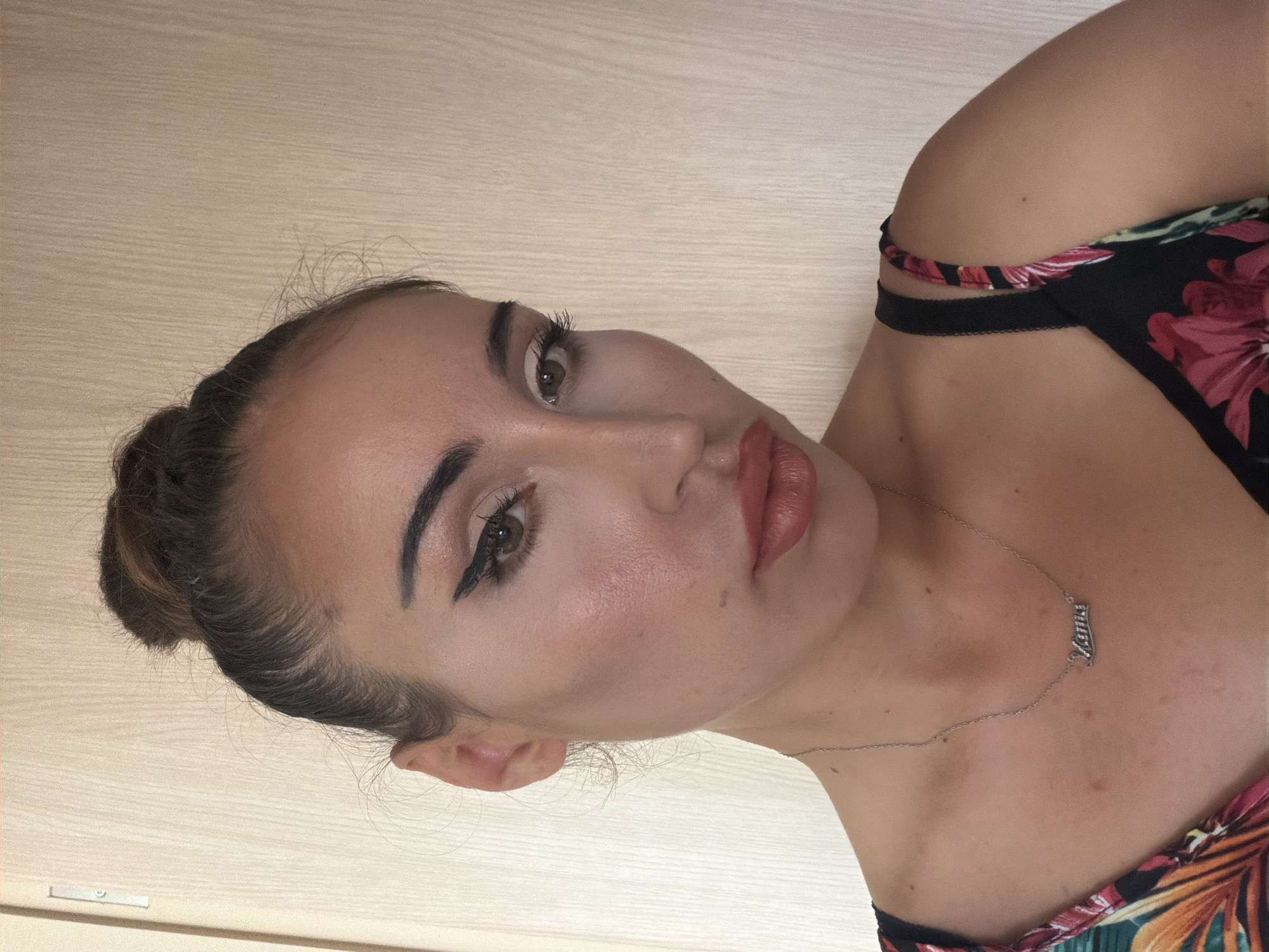 A woman with her hair in a bun is looking towards the camera, her face is partially obscured by the edge of the frame. She is wearing a floral-patterned top and a silver necklace with the word "Mama" spelled out in cursive. The background is a light-colored wall with vertical lines.