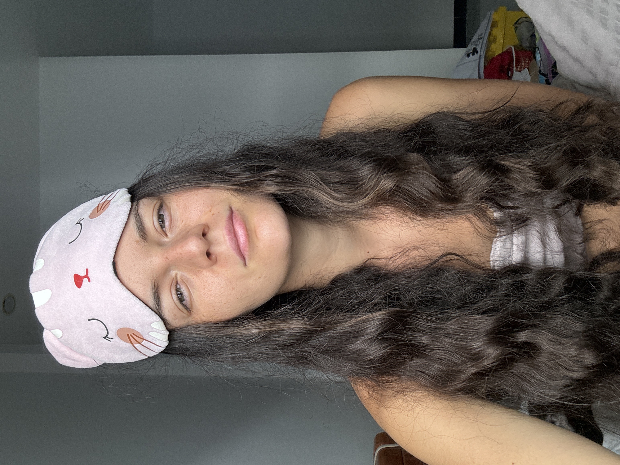 A medium shot depicts a young woman wearing a pink sleep mask with a cat design lying down with her head on a pillow. She has long, dark, wavy hair and appears to be indoors, possibly in a bedroom. The background is nondescript, with a wall and items on a shelf partially visible. The location is not discernable as Mũi Né, Vietnam, without additional details.