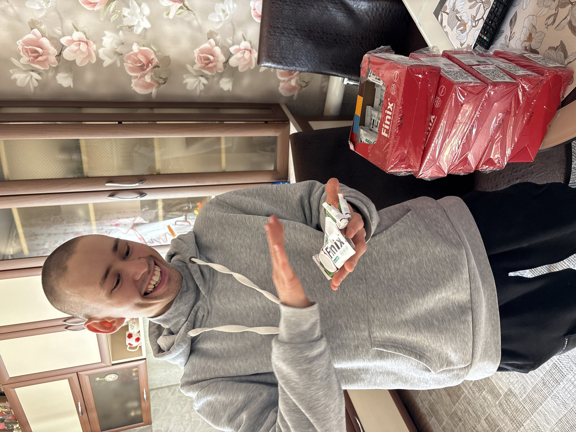 A man with a shaved head, wearing a gray hoodie and black pants, is smiling and holding a "Finix" brand protein bar. He appears to be indoors, with a wooden cabinet and floral wallpaper visible in the background. Several unopened boxes of "Finix" bars are stacked near him on a chair. The image was likely taken in Ulyanovsk, Russia, as the "Finix" brand is associated with that city.