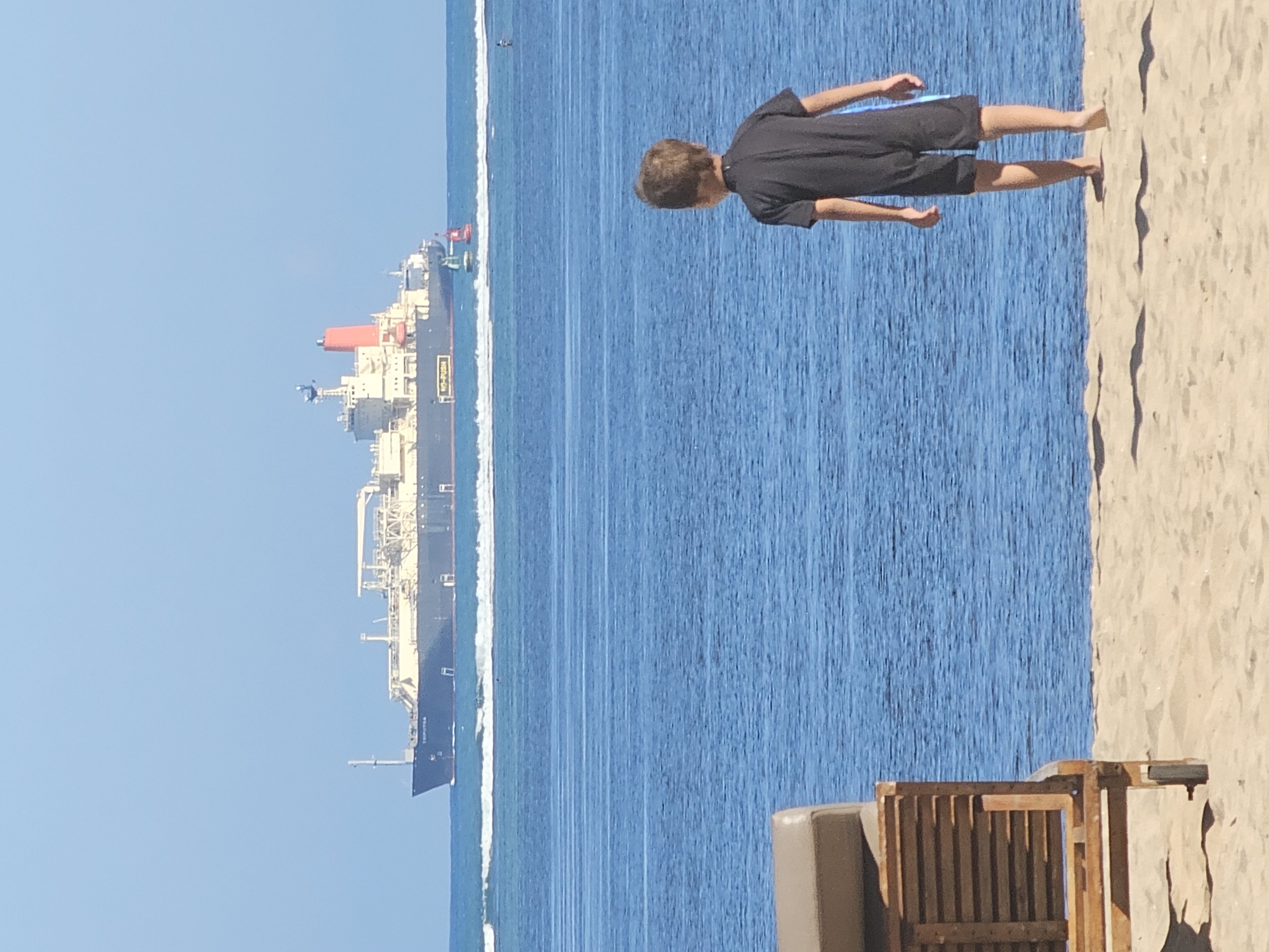 This media file captures a young boy standing on a sandy beach, facing away from the camera and looking out at a large cargo ship on the ocean. The boy is wearing a black t-shirt and dark shorts. The beach sand is a light tan color, and it appears to be a sunny day with clear blue skies.
The ocean is a deep blue, with white waves crashing along the shore. The cargo ship is a prominent feature, appearing to be a liquified natural gas (LNG) carrier, with a long hull and a distinctive superstructure. The ship has the name "TRIPURTI" visible on its side in white lettering.
The scene suggests a relaxed day at the beach, with the boy seemingly captivated by the imposing presence of the ship. The setting appears to be a coastal area, potentially a popular beach destination given the presence of beach furniture in the foreground, including what looks like a wooden chair. The overall mood is one of quiet observation and a sense of scale between the child and the large vessel. The time of day appears to be midday, indicated by the bright sunlight and the shadows cast on the sand.