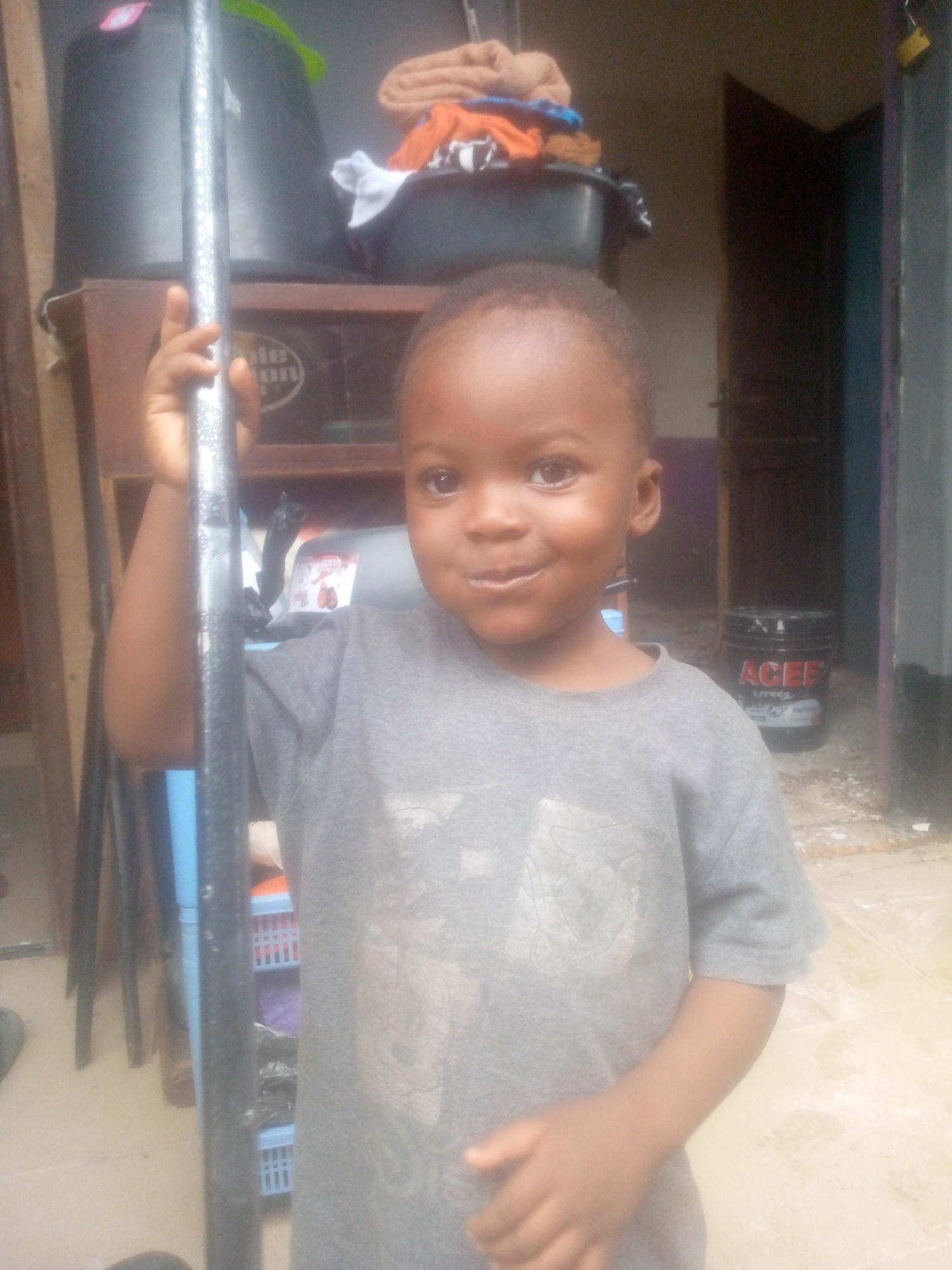 A young child with dark skin, short hair, and a grey t-shirt is holding onto a metal pole in an indoor or partially covered space. Behind the child, there's a wooden cabinet stacked with items, including a black bucket, a container with the word "ole" visible, and a basin holding laundry. A door is slightly open in the background, next to which is a black bucket with "ACEE" printed on it. The floor appears to be tiled. The location is in the city of Unknown, Unknown.