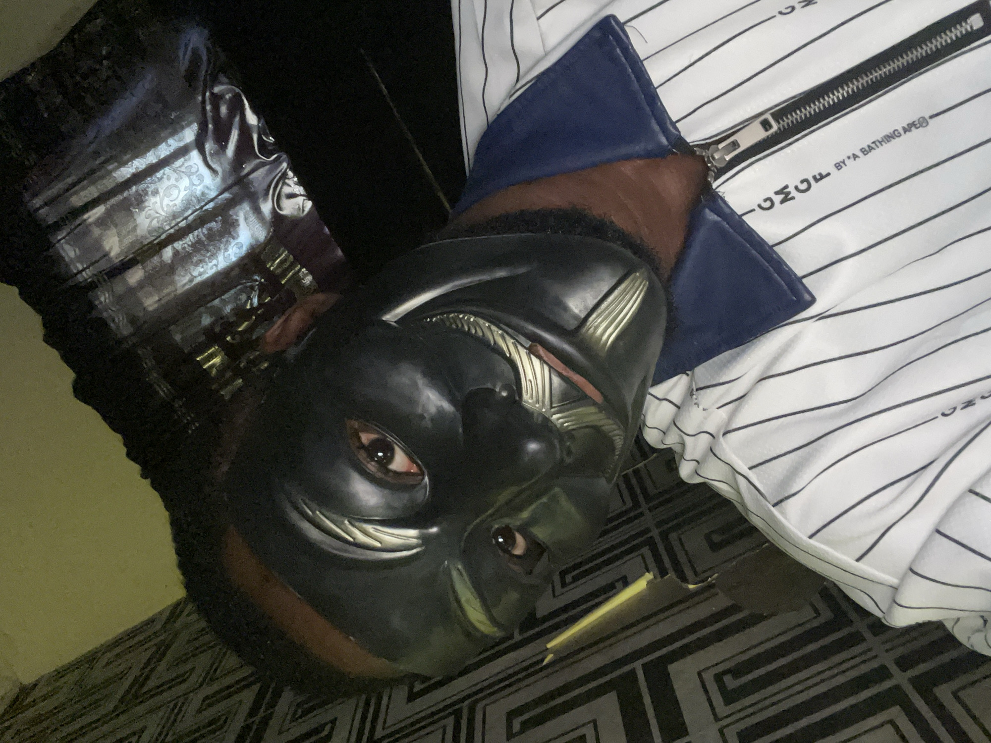 A man wearing a black and silver mask is lying on a patterned floor. He is wearing a white and navy pinstriped jacket with a zipper. The location is not discernible as Lokoja, Nigeria.