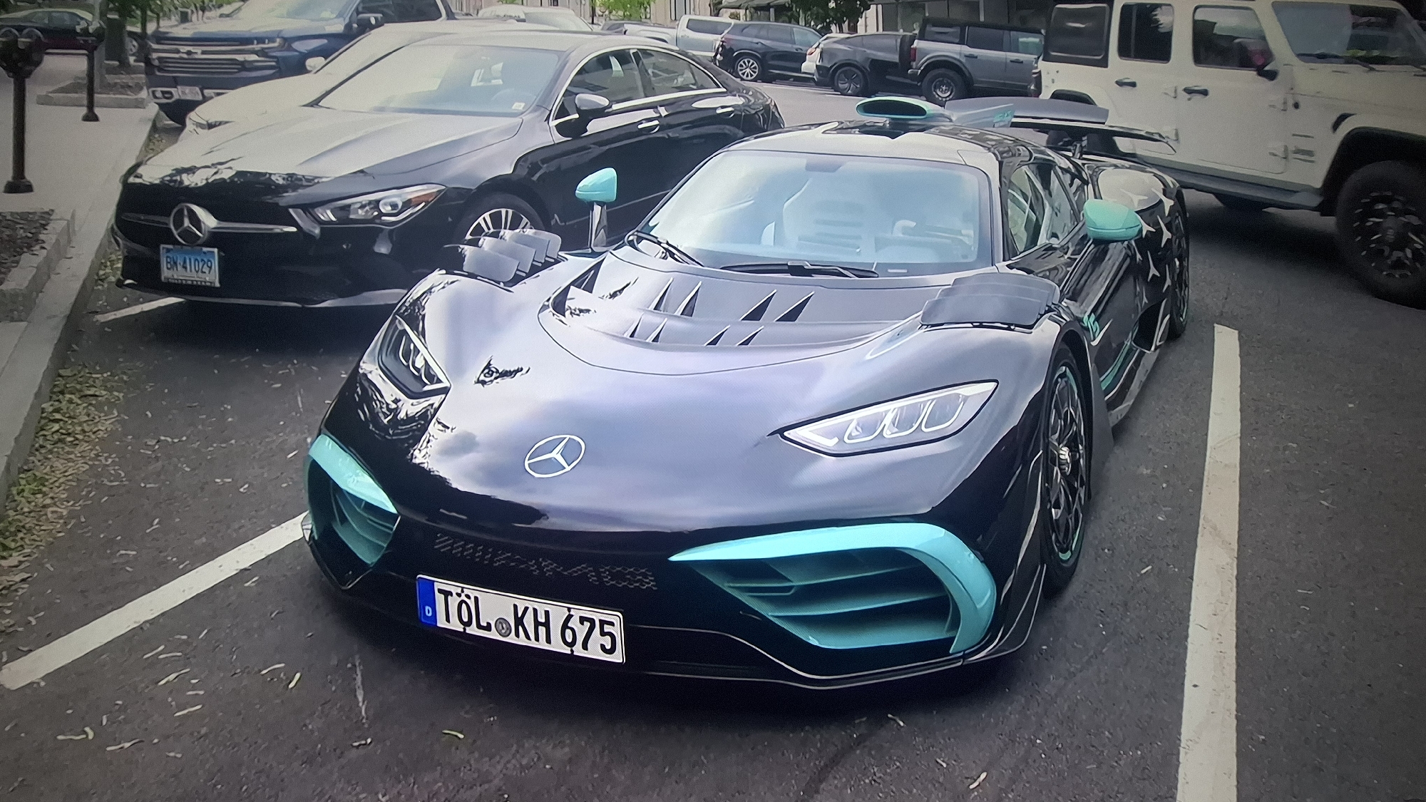 The image shows a black Mercedes-AMG One parked in a parking space on a paved street in an urban environment. The car features turquoise accents. It has a German license plate that reads "TÖL•KH 675". Partially visible in the background are other vehicles, including a black Mercedes sedan with license plate "BN•41029", a gray pickup truck, and a white Jeep. A street lamp post is also visible on the left side of the image. The location is in the city of Unknown, Unknown.