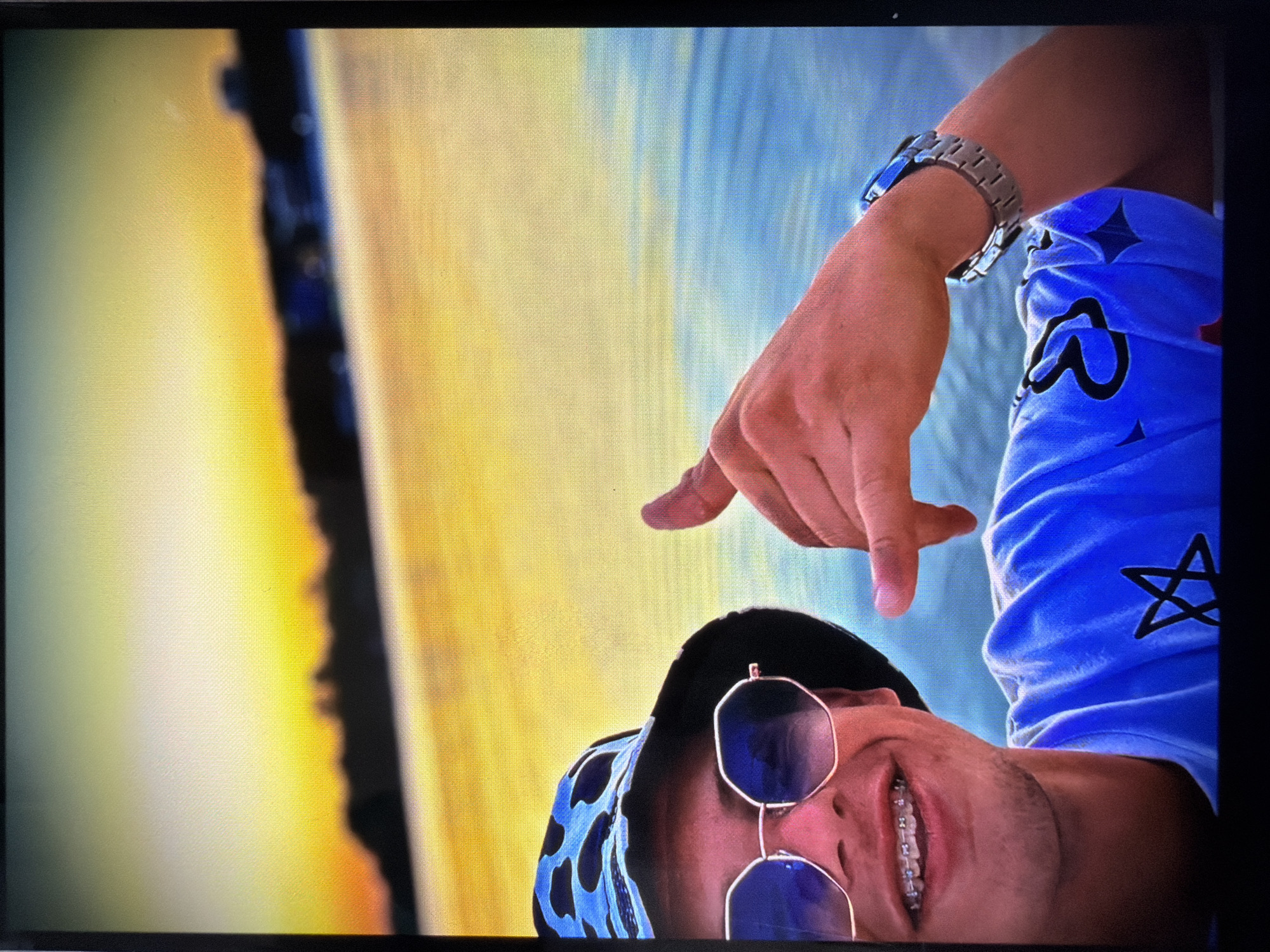 The image, appearing rotated 90 degrees clockwise, presents a close-up portrait of an individual smiling brightly in an outdoor setting. The person, likely male, is positioned in the lower right of the frame (when upright), with their head tilted. They wear octagonal-framed sunglasses with dark lenses and a black cap. A broad smile reveals dental braces.
The individual is dressed in a light blue top featuring black patterns, including stars and other abstract shapes. A silver-toned metal watch with a link bracelet is visible on their left wrist. Their right hand is raised, with the index finger and thumb pointing downwards, possibly in a gesture or pose.
The background is characterized by two distinct horizontal color bands. The upper portion is a vibrant blue, reminiscent of water or sky, while the lower section is a bright, warm gradient of yellow and orange, suggesting sand, land, or strong sunlight. A dark, wavy line separates these two areas, indicating a shoreline, horizon, or the edge of a physical feature. The warm colors strongly suggest the time of day is either sunrise or sunset. The scene evokes a sunny outdoor environment, potentially near a body of water or a resort area, consistent with a location like Razlog, Bulgaria. The person's visible smile conveys happiness or enjoyment. There is no discernible text within the image.
