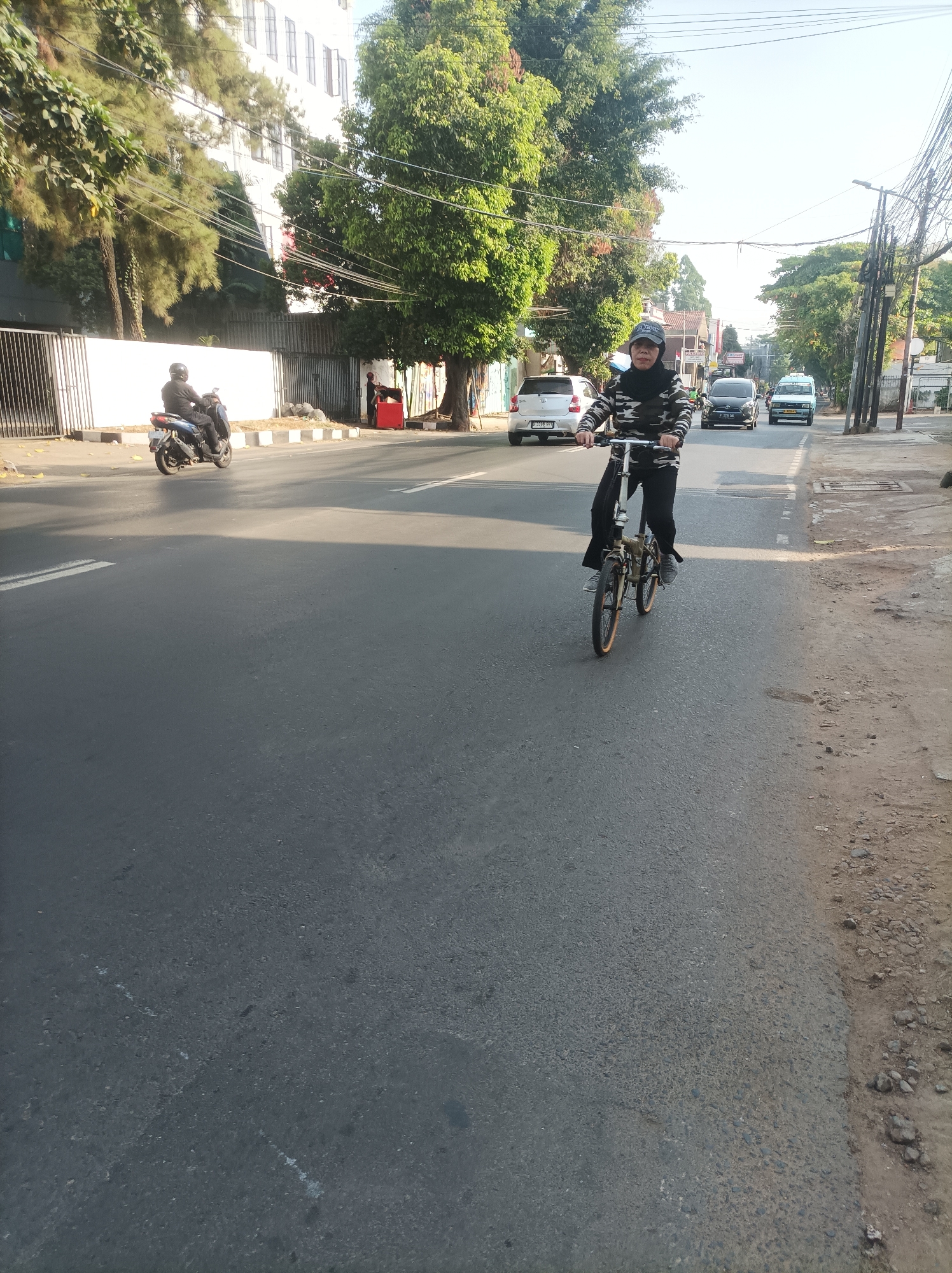 A person wearing a camouflage-patterned jacket and a black headscarf rides a small bicycle on the right side of an asphalt road. The cyclist is in motion, with their hands on the handlebars and feet on the pedals. Several vehicles, including a white car, a black car, and a light blue van, are visible on the road ahead of the cyclist. On the left side of the road, a person on a scooter is seen passing by. In the background, there is a multi-story white building with several windows, surrounded by lush green trees. Power lines and street lights are also present. The scene appears to be a street in South Jakarta, Indonesia, with typical urban infrastructure and vegetation. The time of day seems to be either early morning or late afternoon, given the lighting and shadows.