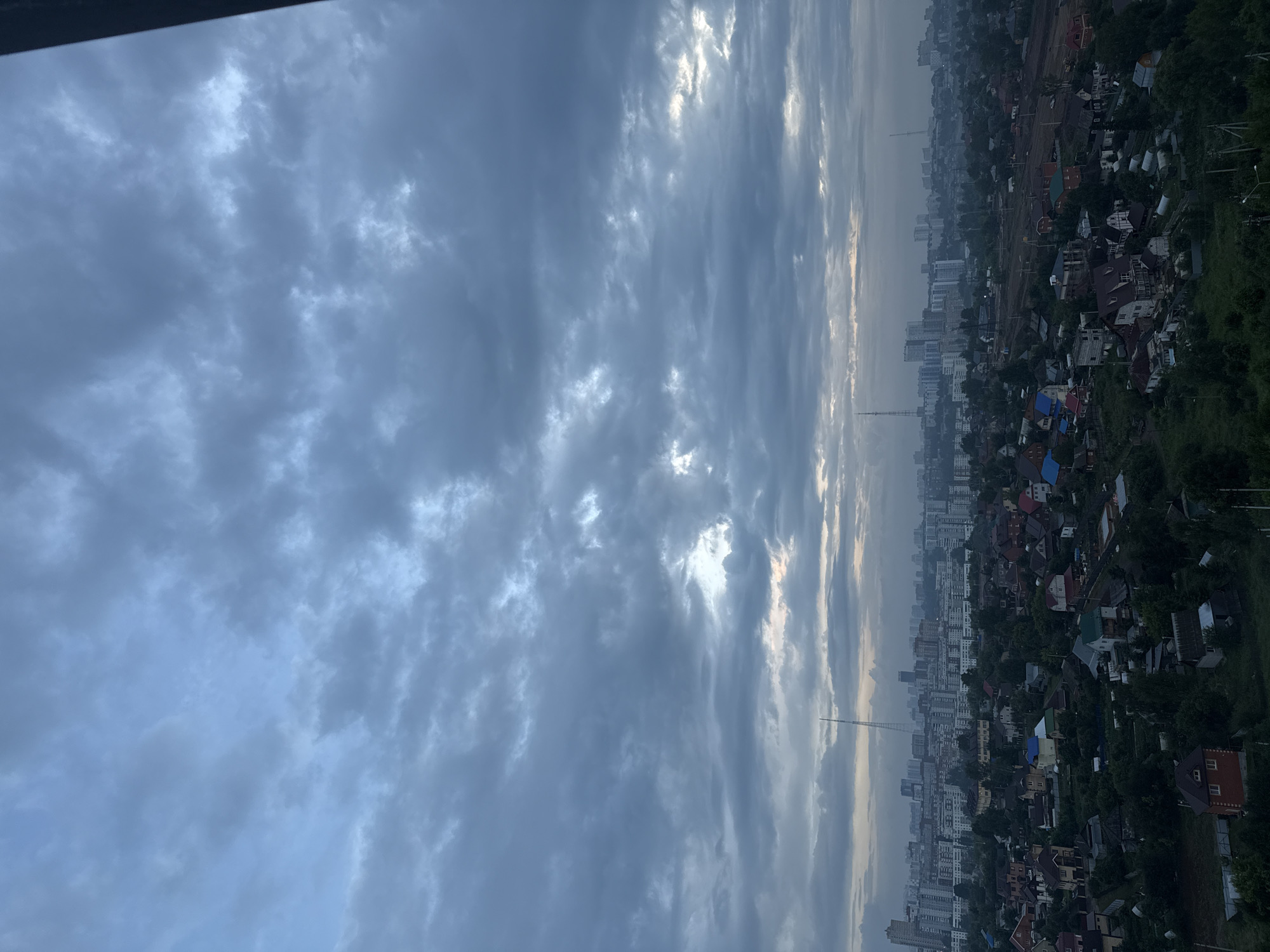 The image depicts an aerial view of a city, likely Perm, Russia, with a strong focus on the sky and clouds. The scene is oriented vertically, suggesting it was taken from a high vantage point, possibly a building or drone.
The upper portion of the image is dominated by a dramatic sky filled with a mixture of dark, heavy clouds and brighter, more diffused patches where sunlight is breaking through. These clouds create a sense of depth and texture, with some appearing wispy and others more solid and ominous. The lighting suggests it might be early morning or late evening, as the sun is low on the horizon, casting a soft, diffused light.
Below the sky, the landscape of a city unfolds. A dense collection of buildings, ranging from multi-story apartment blocks to smaller houses with pitched roofs, stretches across the frame. The architecture appears to be a mix of modern and older styles, with varying colors of roofs and walls visible. Lush green trees are interspersed among the buildings, indicating a significant amount of vegetation within the urban environment.
A tall, slender tower, possibly a radio or communication mast, is prominently visible in the middle ground, rising above the surrounding structures. The overall impression of the scene is one of a city under a cloudy sky, with a hint of urban sprawl and pockets of green space. There is no visible text in the image. The weather appears to be overcast with some breaks of sunlight, and the time of day is likely during twilight hours. The scene evokes a feeling of vastness and the interplay between natural elements like the sky and the man-made environment of the city.