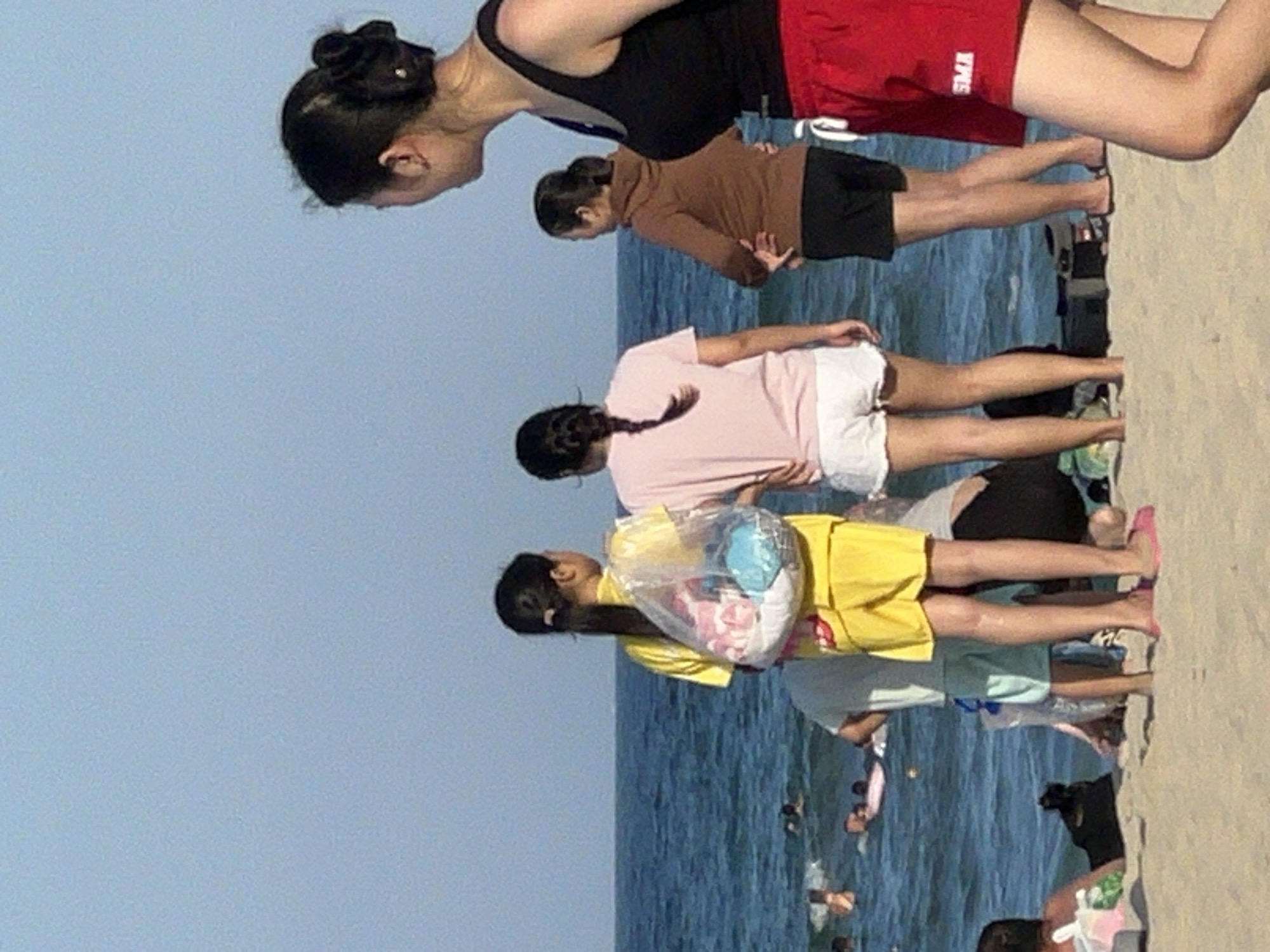 The image shows a group of people on a beach, likely in the city of Đà Nẵng, Vietnam, given the prevalence of beaches there. In the foreground, a woman in a black tank top and red shorts stands facing away from the camera, her hair pulled back. To her left, another woman wearing a brown hoodie and black shorts is also facing away. Further to the left, two young women stand side-by-side. One is wearing a yellow top and shorts, carrying a clear plastic bag filled with colorful items. The other is wearing a pink t-shirt and white shorts. In the background, the ocean is visible with several people swimming or wading in the water. The scene appears to be a typical day at the beach with many people enjoying the water and sand. The sky is clear and blue, indicating good weather.