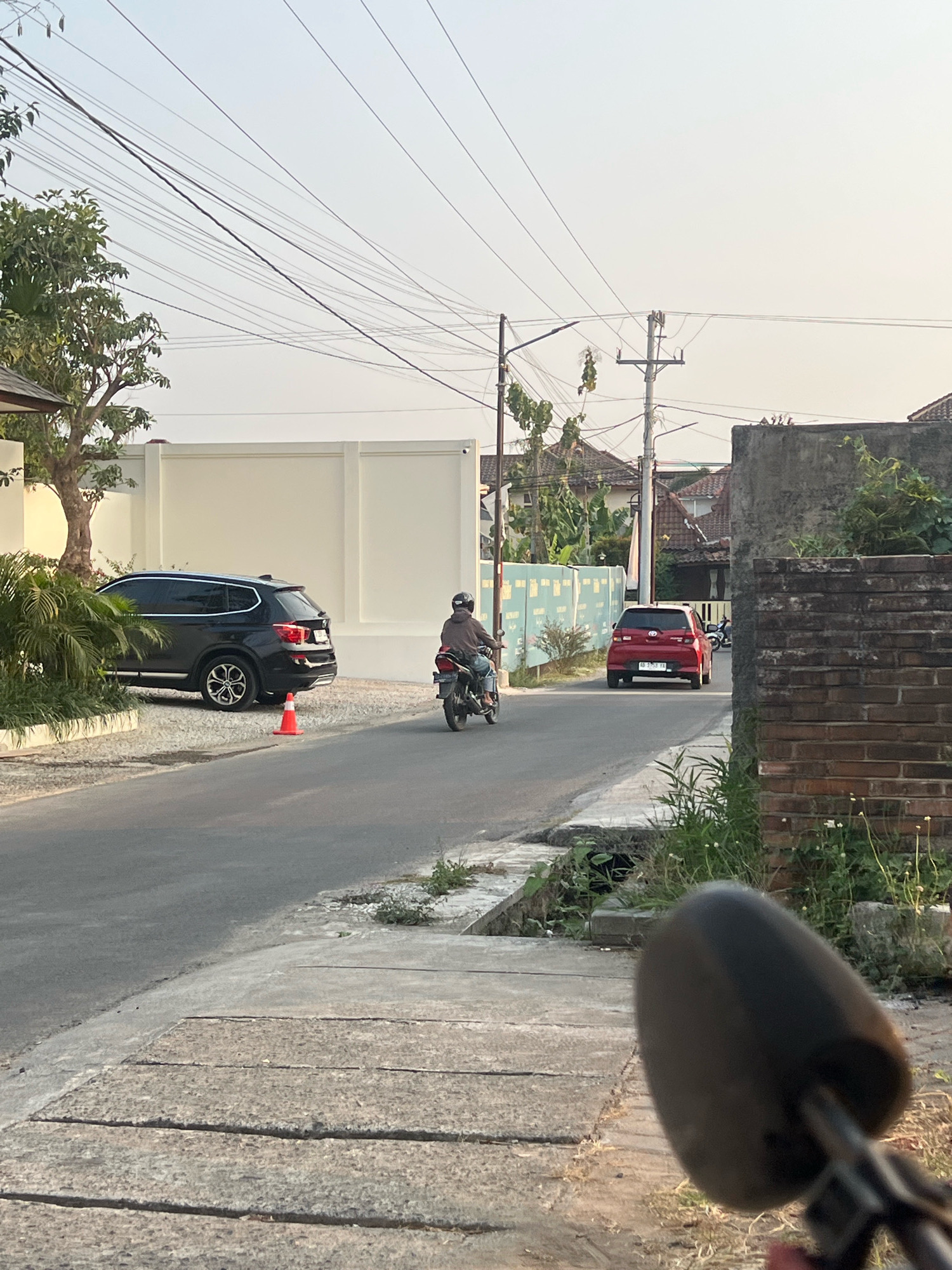 The image shows a street scene in Ngaglik, Indonesia. A person on a motorcycle is riding down the street, heading away from the camera. The motorcycle is dark-colored, and the rider is wearing a brown jacket and a helmet. To the left of the motorcycle, there is a black SUV parked in a driveway. The SUV is a BMW X5. In front of the SUV, there is an orange traffic cone.
The street is paved with asphalt, and there is a concrete sidewalk on the right side of the road. There are power lines running overhead, supported by utility poles. Buildings line the street, including a white wall with a blue patterned fence in front of it. To the right of the street, there is a brick wall and overgrown vegetation. The sky is clear and bright, suggesting it is daytime. The overall impression is that of a quiet residential street in a suburban area.