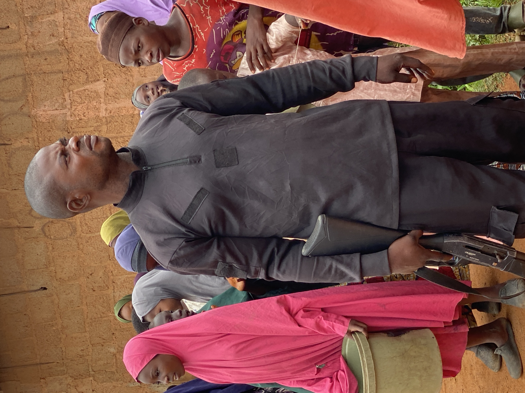 In Kawo, Nigeria, a group of people, predominantly women and children, are gathered outdoors in front of a rough-textured, mud-brick wall. A man in the foreground, wearing a black zipped shirt and pants, holds a shotgun in his right hand. He has a serious expression and is looking to his right. To his left, a young girl in a bright pink hijab holds a light green bucket. Behind the man, several other individuals are visible, including a young person wearing a brown knitted cap and a red patterned shirt, and others with hijabs in various colors like purple, green, and yellow. The ground appears to be dirt.