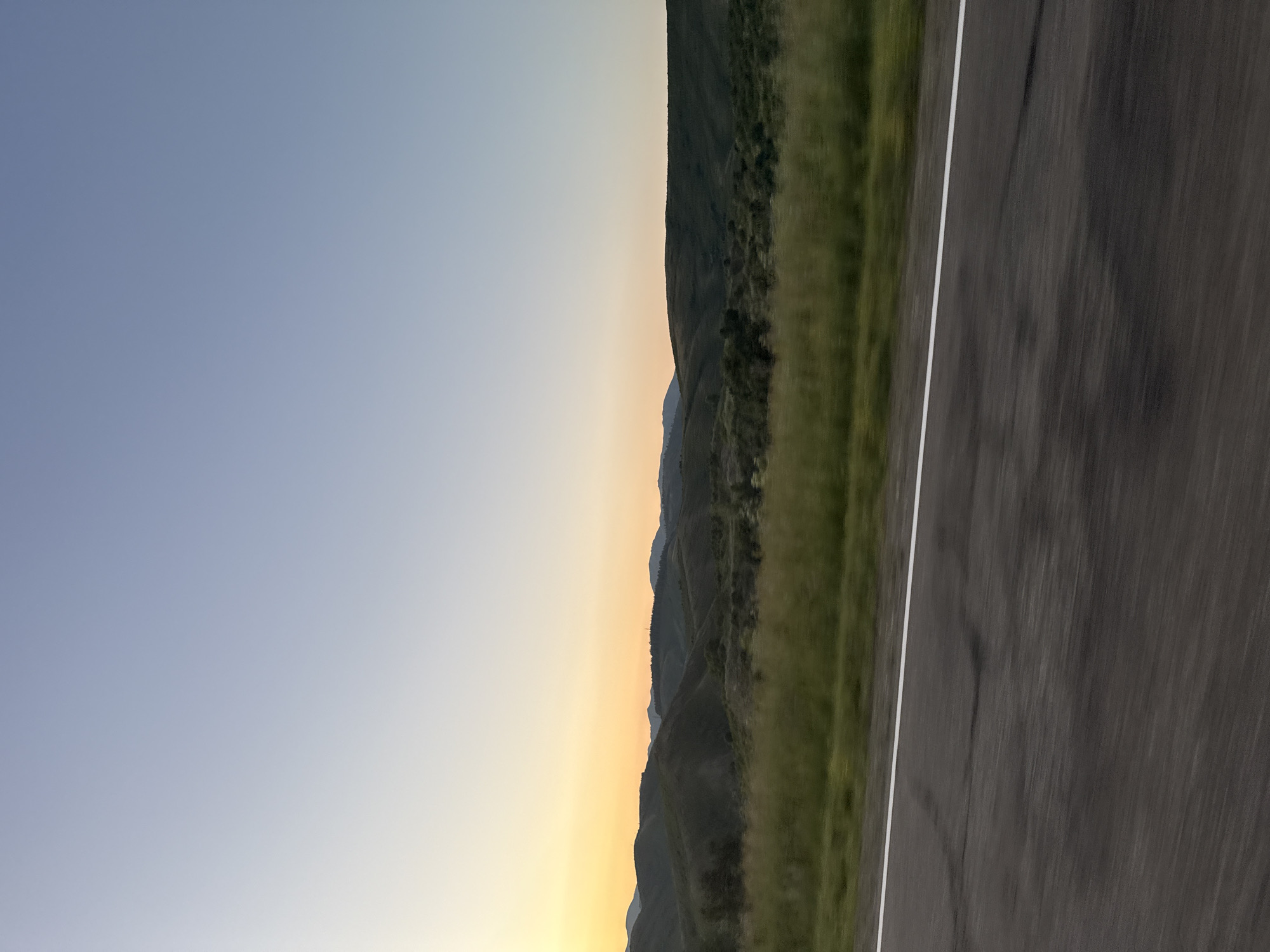 The image shows a wide, panoramic view of a rural landscape with rolling hills under a clear, gradient sky that transitions from pale yellow near the horizon to a soft blue overhead. The sun is low, casting a warm glow on the distant hills, which are covered in dark green vegetation. In the foreground, a paved road with a white line marking the lane stretches across the frame, showing motion blur suggesting it was taken from a moving vehicle. To the left of the road, a strip of blurred green grass and vegetation runs along the roadside. The overall impression is one of a serene, undeveloped natural environment. This scene is consistent with the rural surroundings of Severnoe, Kazakhstan, which features open landscapes and agricultural land.