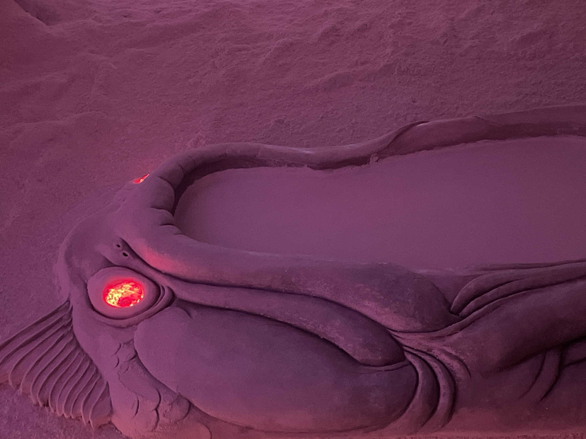The image depicts a large snow sculpture of a fish-like creature with exaggerated features, possibly a sculpture of a lamprey. The sculpture has a prominent eye that is illuminated with a red light. The sculpture also has what appears to be a trough carved into the back of it. The whole scene is bathed in magenta light. While the image doesn't explicitly state the location, the prompt mentions Vinnytsia, Ukraine as the potential location.