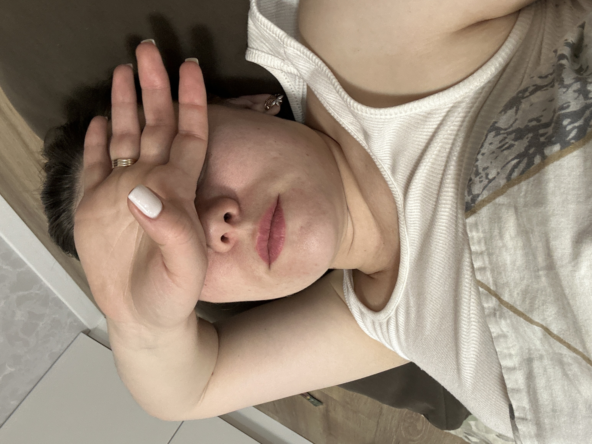 A woman lies on her side, shielding part of her face with her hand. She has short, dark hair, fair skin, and wears a white tank top. A ring is visible on her hand. The setting appears to be indoors, with a light-colored surface on the left and a darker surface behind her head. No information suggests this image was taken in Chaikovsky, Russia.