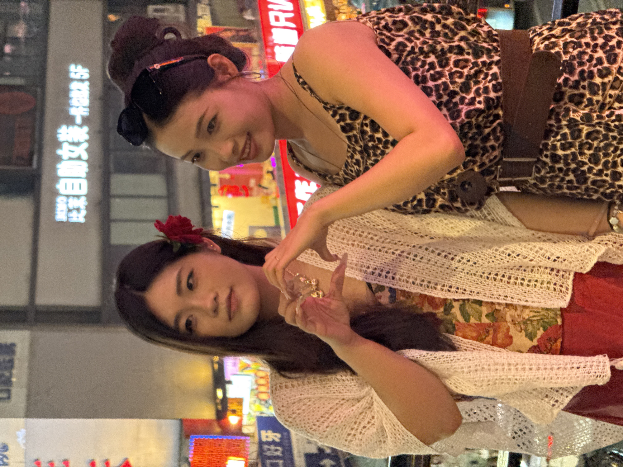 The image features two young Asian women posing in an urban environment at night. The background is a vibrant blur of colorful neon signs and illuminated buildings, suggesting a lively city street, consistent with the context of Liangjiang, China.
The woman positioned higher in the frame has dark hair styled up with sunglasses resting on her head. She wears a leopard-print spaghetti strap dress or top, cinched with a wide, dark brown belt. She smiles warmly, looking towards the second woman.
The second woman, positioned lower, has long, dark hair adorned with a red flower behind her ear. She is wearing a white, open-knit crochet cardigan over a floral-print top, with a red garment (possibly a skirt or pants) visible underneath. Her hands are forming a heart shape, and she is holding a piece of golden jewelry, possibly a necklace or bracelet. She looks towards the viewer. She also wears rings and a bracelet.
A prominent, brightly lit sign in the background displays Chinese characters: "白丽人女装 - 鞋业部" (Bailiren Women's Wear - Footwear Department). Other less distinct Chinese characters are visible on surrounding signs. The setting is clearly nighttime due to the reliance on artificial lighting. Both women appear cheerful and engaged in a friendly interaction.