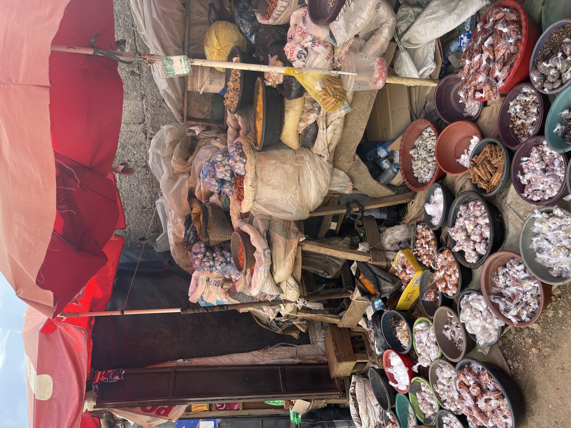 This image depicts an outdoor market stall in Rafin Par, Nigeria, featuring a variety of goods, primarily food items and spices, displayed under a large red and white umbrella. The stall is cluttered with numerous bowls and sacks filled with produce. In the foreground, several dark-colored bowls are filled with small, individually wrapped items, likely spices or dried goods. To the left, a collection of garlic bulbs is arranged in a brown bowl. Moving towards the back, more bowls contain similar packaged items. Behind these, large sacks are piled high, some containing what appear to be onions and chili peppers. A yellow bag of what could be rice or grain is visible, along with larger sacks of various commodities. The stall structure is made of rough wooden planks, and the background shows a rough, possibly concrete or brick wall. A thin white pole supports the umbrella. No individuals are clearly visible in the scene, though the setting suggests a commercial activity. The ground is dirt and appears uneven.