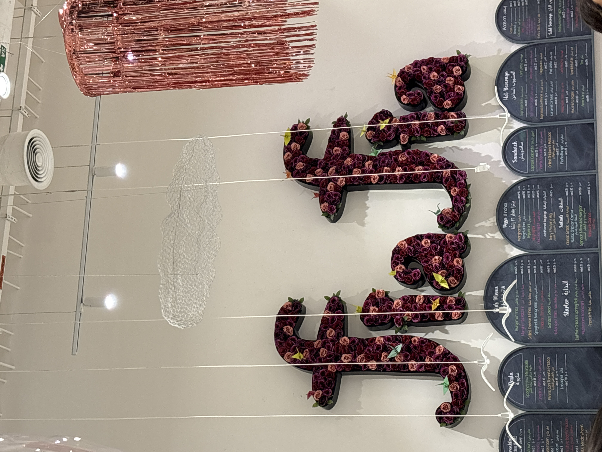 The image shows the interior of a restaurant in Dubai, United Arab Emirates. The wall is decorated with large, floral "f" letters, suggesting the restaurant's name might be related to "fafa." Below the floral letters, there are several chalkboards displaying menus. The chalkboards are organized into sections like "Kids Menu," "Starter," "Pizza 12 inches," "Sandwich," "Hot Beverage," and "Cold Beverage," with items listed in both English and Arabic. The restaurant appears to have a modern and chic ambiance, with hanging decorative elements such as clouds and small paper birds. There are no people visible in the image.