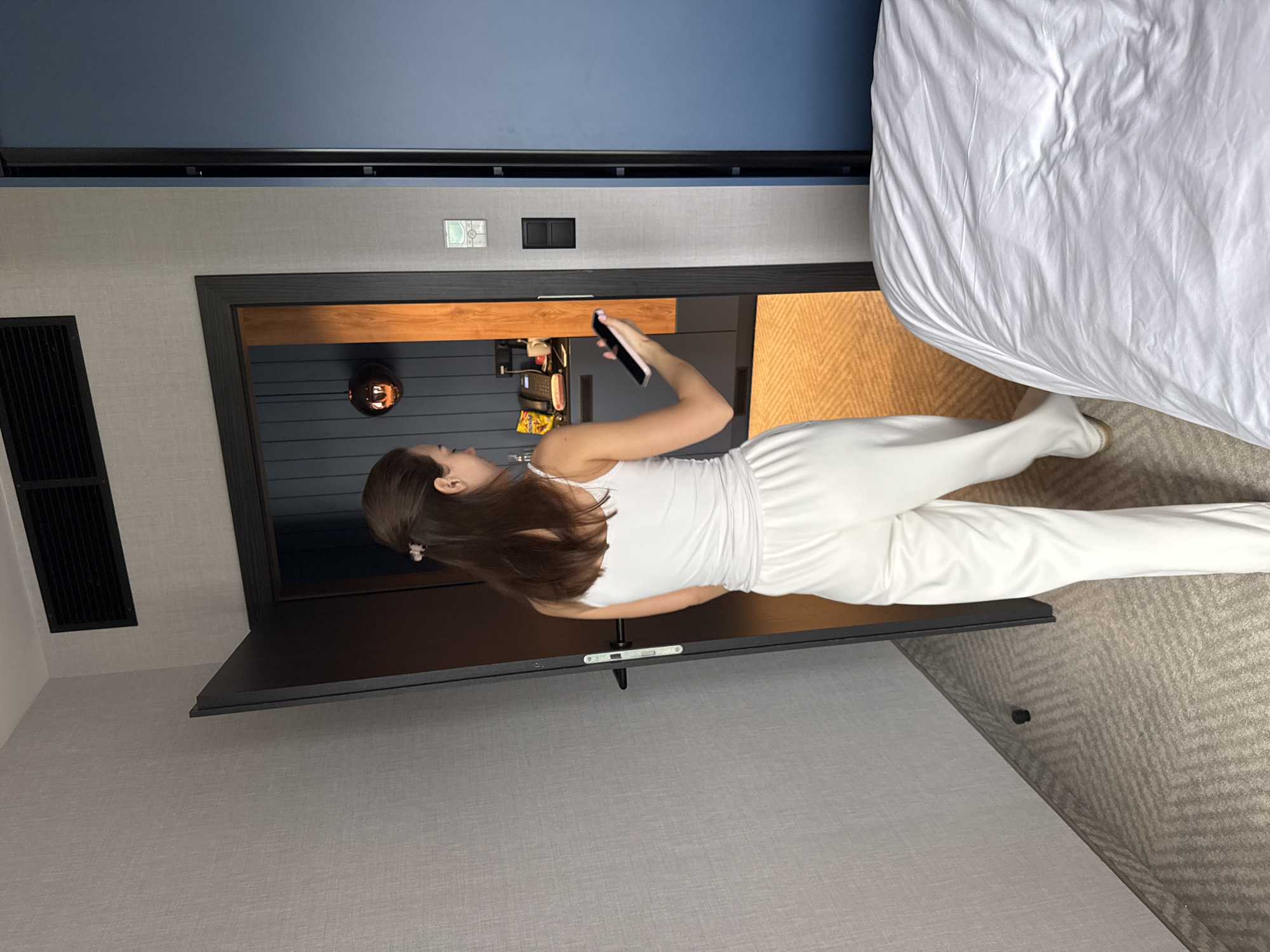 The image captures a young woman standing in what appears to be a modern hotel room or bedroom. She has long, dark brown hair and is wearing a white tank top and matching white pants. She holds a black smartphone in her right hand, seemingly using it.
Directly in front of her is a large, dark-screened television, likely off, mounted on a wall. Below the TV, a wooden shelf or cabinet runs along the wall, featuring a spherical decorative light and what looks like bags and snacks. The wall behind the woman and the TV is composed of a lower textured light grey section and an upper dark blue section. Above this, a lighter blue ceiling is visible.
To the woman's right, a bed with white, rumpled sheets occupies a significant portion of the frame. The floor beneath her and extending throughout the room is covered with a light-colored carpet featuring a herringbone pattern. To her left, on the grey wall, a black ventilation grille is visible. On the grey wall above the wooden shelf, there are black electrical outlets and a small white square control panel, possibly a thermostat. The overall setting suggests a contemporary and comfortable indoor environment. No visible text is clearly discernible.