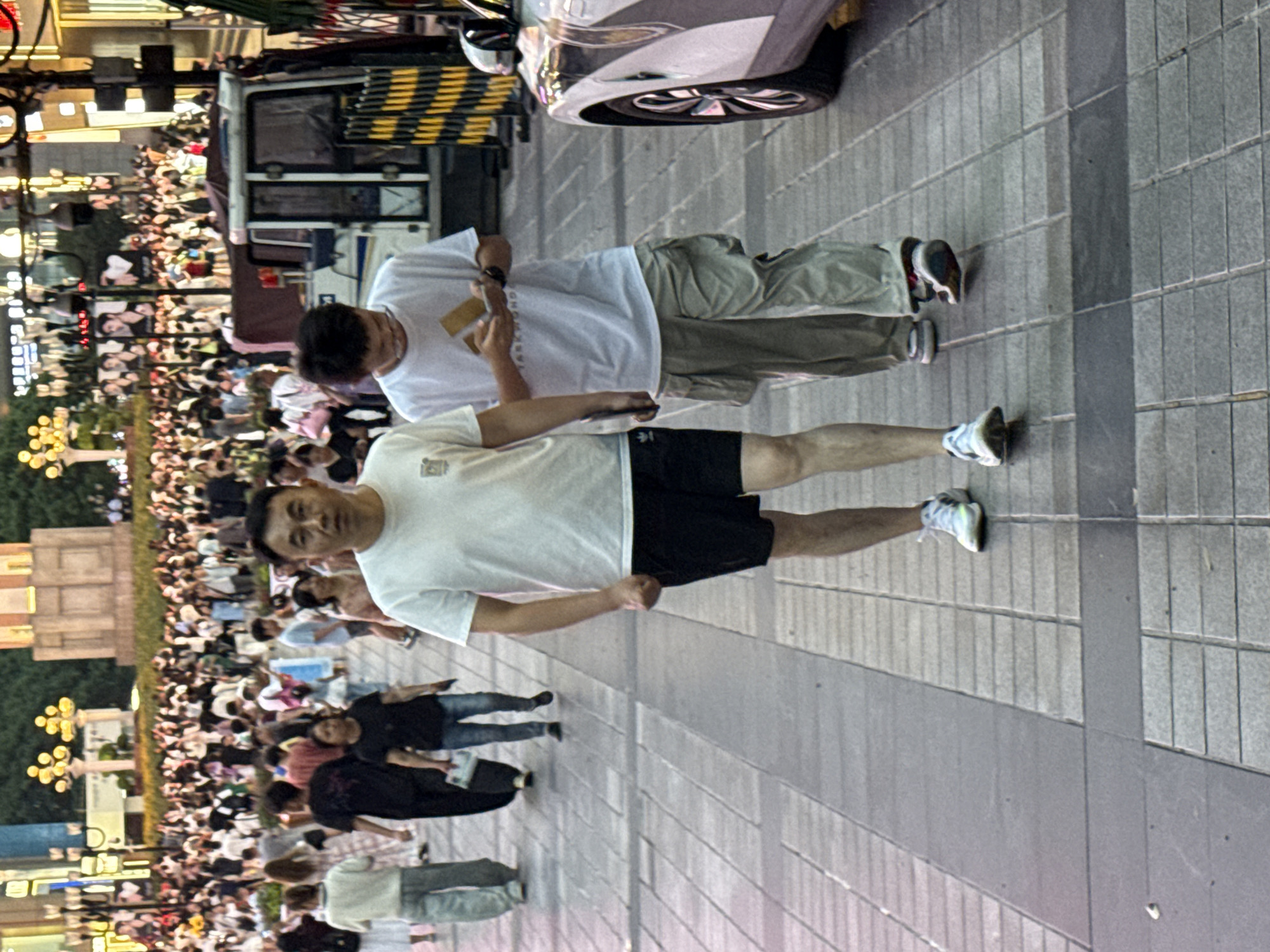 In Yuzhong District, China, two men walk down a pedestrian walkway lined with shops. The man on the left, wearing a white t-shirt and black shorts, walks with his left arm bent at the elbow and his fist clenched. The man on the right, wearing a white t-shirt and khaki cargo pants, looks down at his phone. In the background, a crowd of people walks along the street, and a white vehicle is parked on the right side of the image. The walkway is paved with gray tiles, and streetlights with yellow bulbs are visible in the distance. The scene appears to be a busy commercial area during the day.