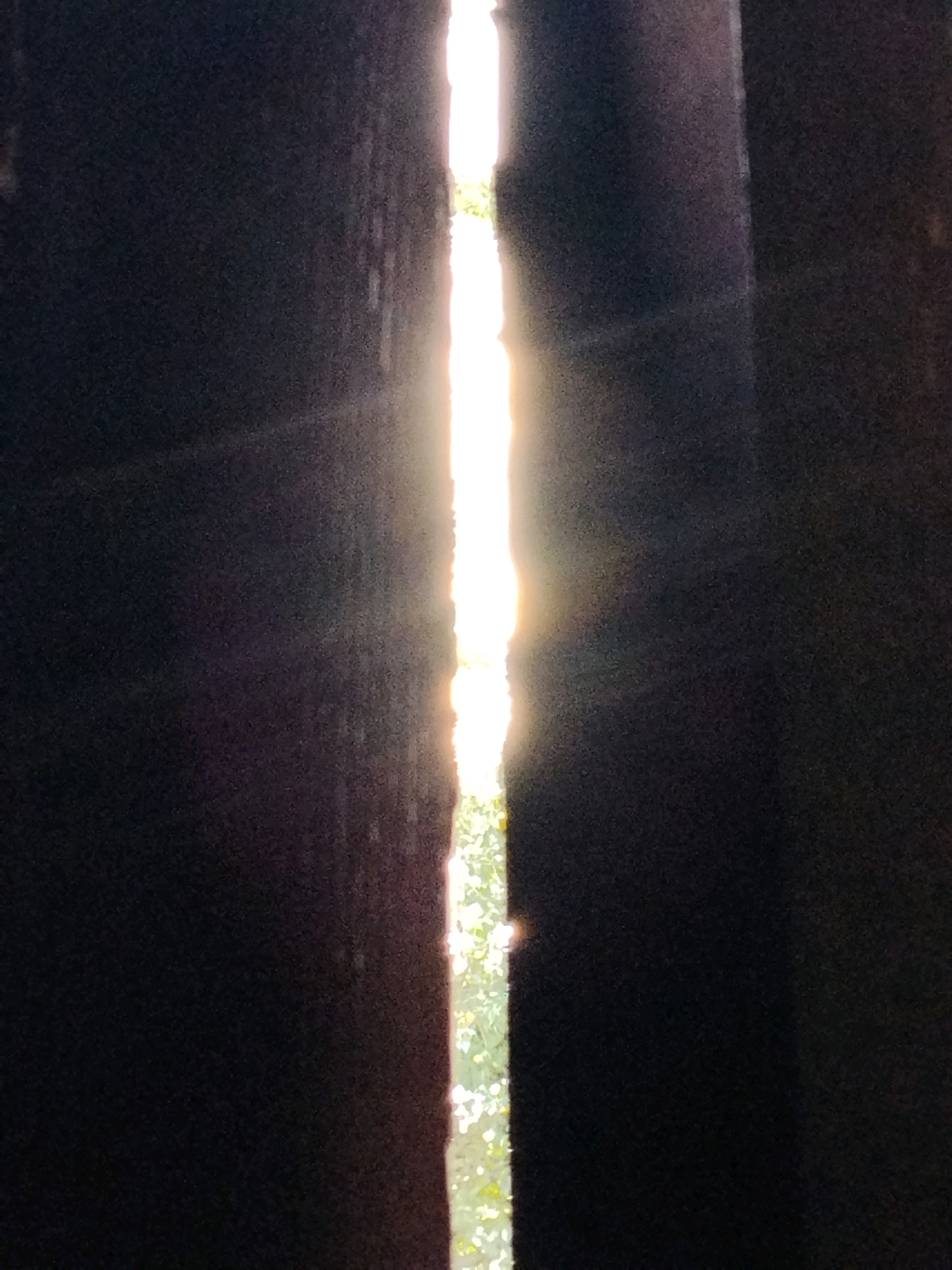 The image depicts a narrow vertical sliver of bright sunlight breaking through a dark opening, possibly a gap between two structures or within a dense forest canopy. The light source is intense and appears to be the sun, with visible rays emanating outwards. The surrounding areas are in deep shadow, with textured surfaces that are difficult to discern clearly. To the left and right of the light, dark, rough textures are visible, suggesting man-made structures or dense foliage. Through the illuminated gap, glimpses of green foliage and possibly trees can be seen in the background. The overall impression is one of light piercing through darkness. Without further context or landmarks, it is impossible to determine the specific location within the city of Hoholeve, Ukraine.