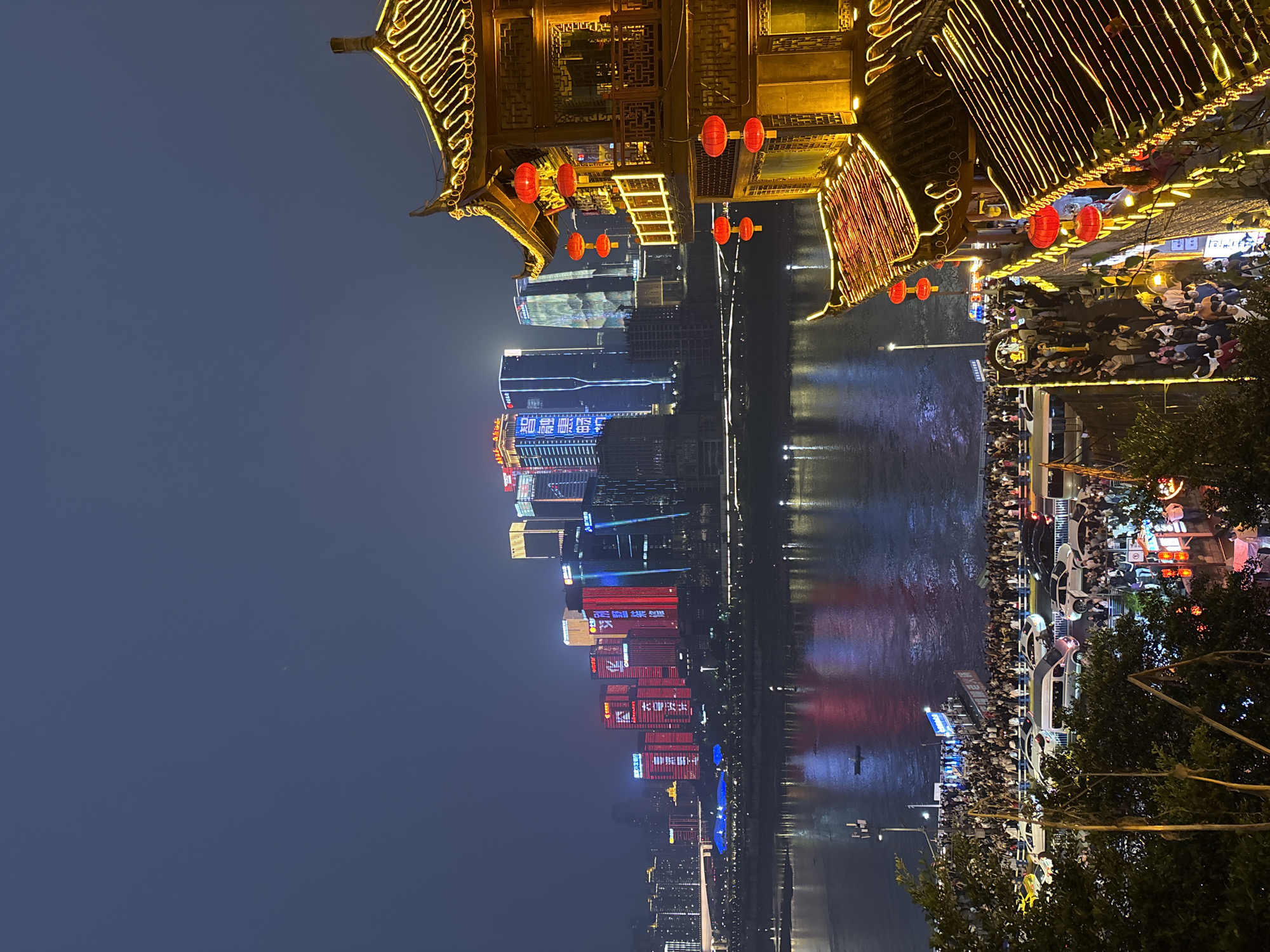 The image captures a nighttime view in Yuzhong District, Chongqing, China. A body of water, likely the Jialing River, reflects the illuminated skyline of modern skyscrapers with colorful displays. Traditional Chinese architecture with ornate roofs, lanterns, and decorative lighting lines the waterfront, with large crowds of people strolling along the promenade. The scene is alive with vibrant colors and the interplay of modern and traditional elements.