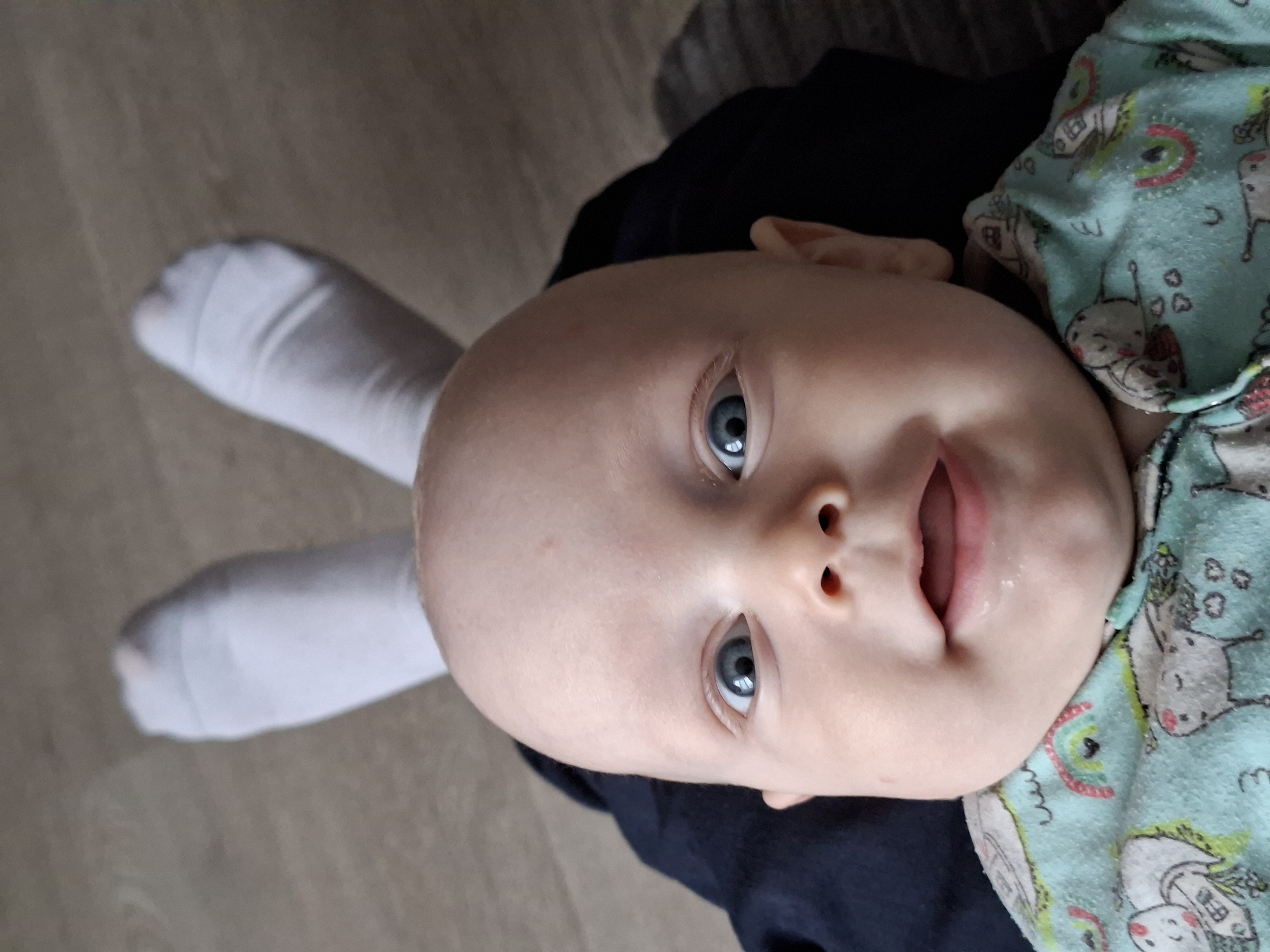 The image shows a baby with blue eyes and a bald head wearing a blue and green patterned onesie. Two white socks are positioned near the baby's head, resembling rabbit ears. The baby appears to be lying down on a dark blue or black fabric. The background consists of a wooden floor and a section of grey fabric. The location is unknown.