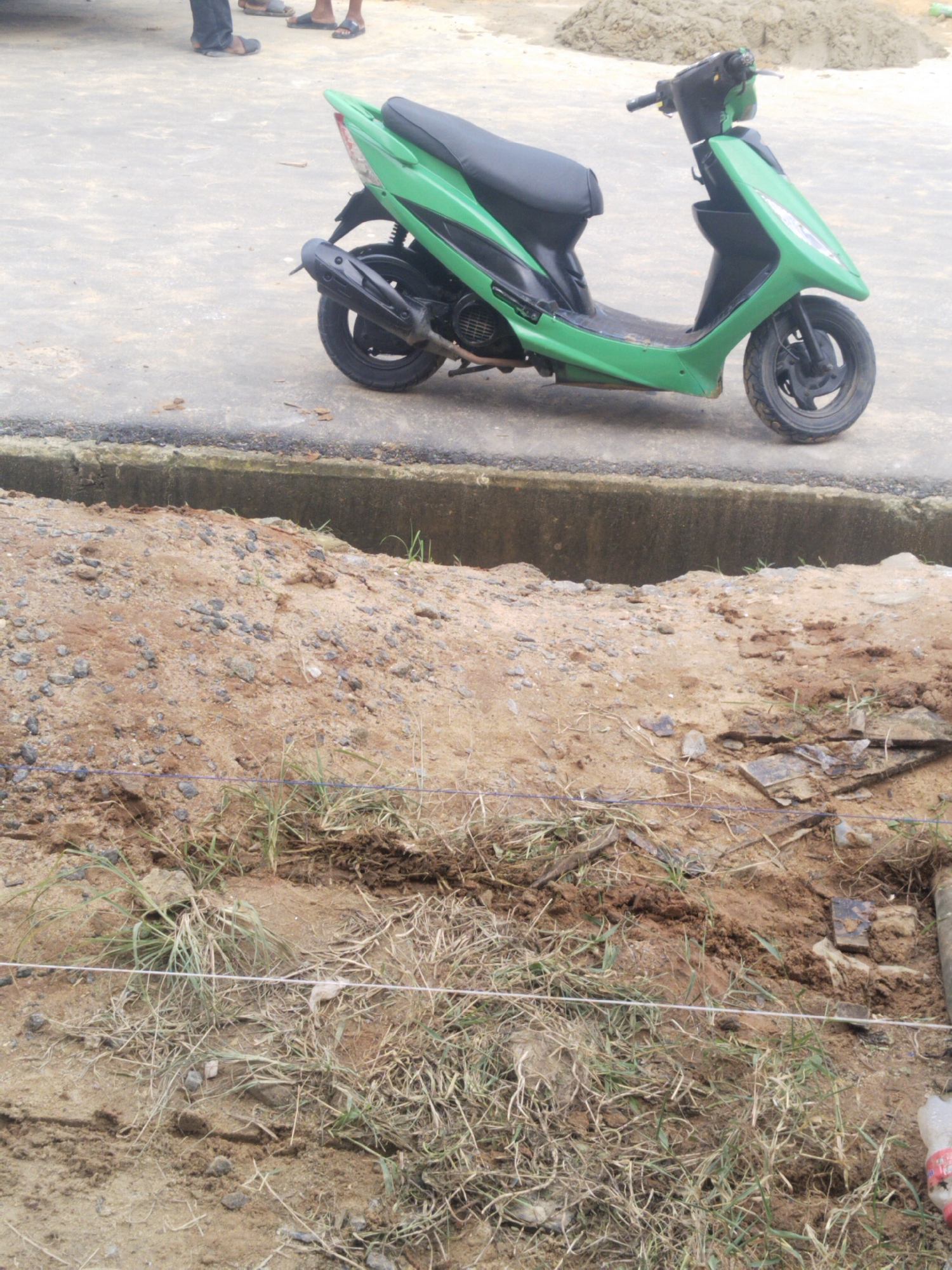 An overhead, slightly angled medium shot captures a green and black scooter parked on a concrete surface in front of a dirt mound. The scooter is positioned with its front wheel facing right and its rear wheel facing left. The concrete area shows some scattered debris. To the right of the scooter, a mound of brown dirt is visible, with some sparse green grass growing on its surface. In the background, beyond the dirt mound, is a pile of sand. The lower portion of the image is dominated by the dirt mound and a more detailed view of the ground, including more grass, loose soil, and a few small rocks. A thin white string is stretched horizontally across the lower part of the frame.