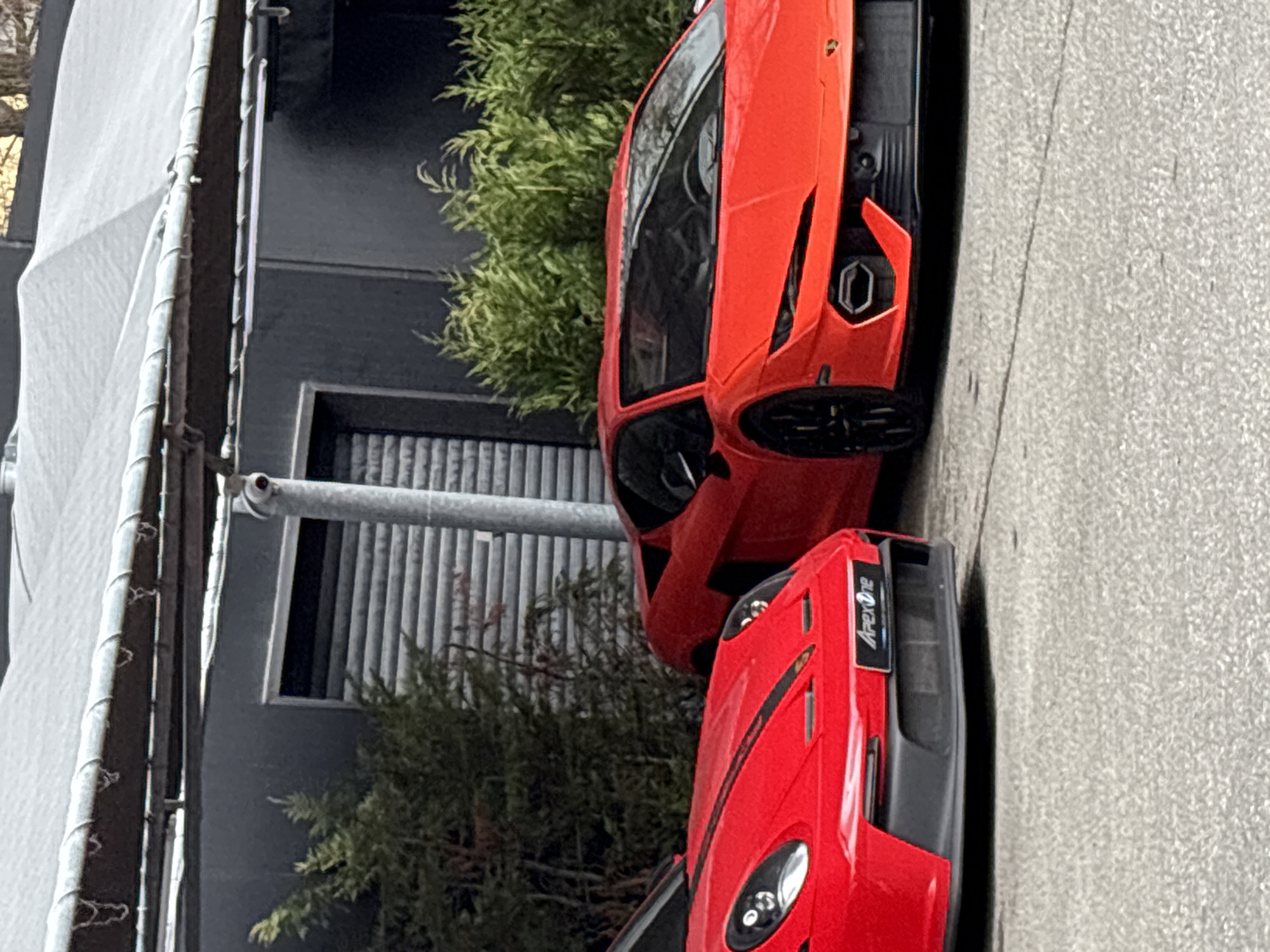 The image shows two parked cars, one orange Lamborghini Huracan and one red car with an "Apex One" logo, on an asphalt road in an urban environment. Behind the cars is a building with a grey facade, a roller shutter window, and some greenery. A scaffolding structure is visible on the left side of the image. The location is possibly in Sofia, Bulgaria, based on general streetscape features, but there is no explicit information to confirm this.