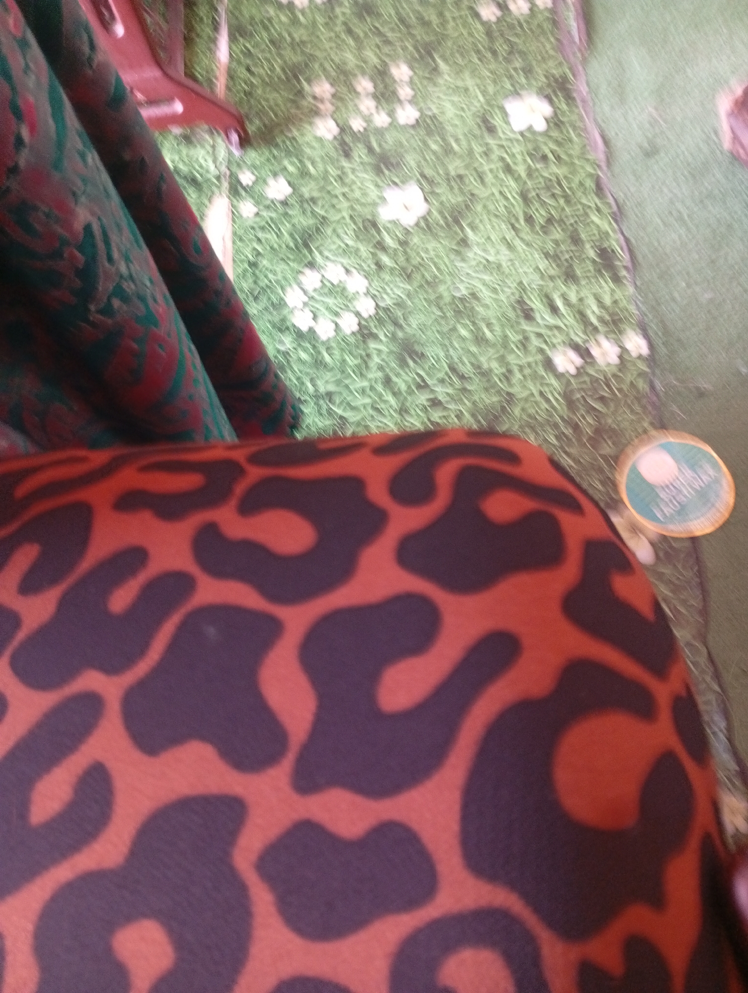 An extreme close-up, angled shot shows a portion of a red and black leopard print fabric filling the foreground. The fabric appears to be a flexible material, possibly clothing or upholstery, with large, irregular dark purple spots on a bright orange-red background. The texture of the fabric is visible, with a slight sheen.
In the background, the scene is out of focus. A green surface with a pattern of white floral shapes is visible, resembling a lawn with flowers, or perhaps a decorative rug or mat. To the right of this, a blurred patch of green indicates another surface, possibly carpet or grass.
On the right side of the background, a circular, metallic object is present. It appears to be a tag or a coaster with a green and white design, and some text is partially visible on it in a light color.
Partially visible in the upper left is a dark red, curved object, possibly part of a chair or a piece of furniture, with a draped fabric showing a pattern of dark green and red paisley designs.
The lighting is diffused, with no harsh shadows. The overall impression is of an indoor setting, perhaps a living room or a hobby area, with a focus on decorative textiles. The specific location in the city of Unknown, Unknown, cannot be determined from the visual information provided.