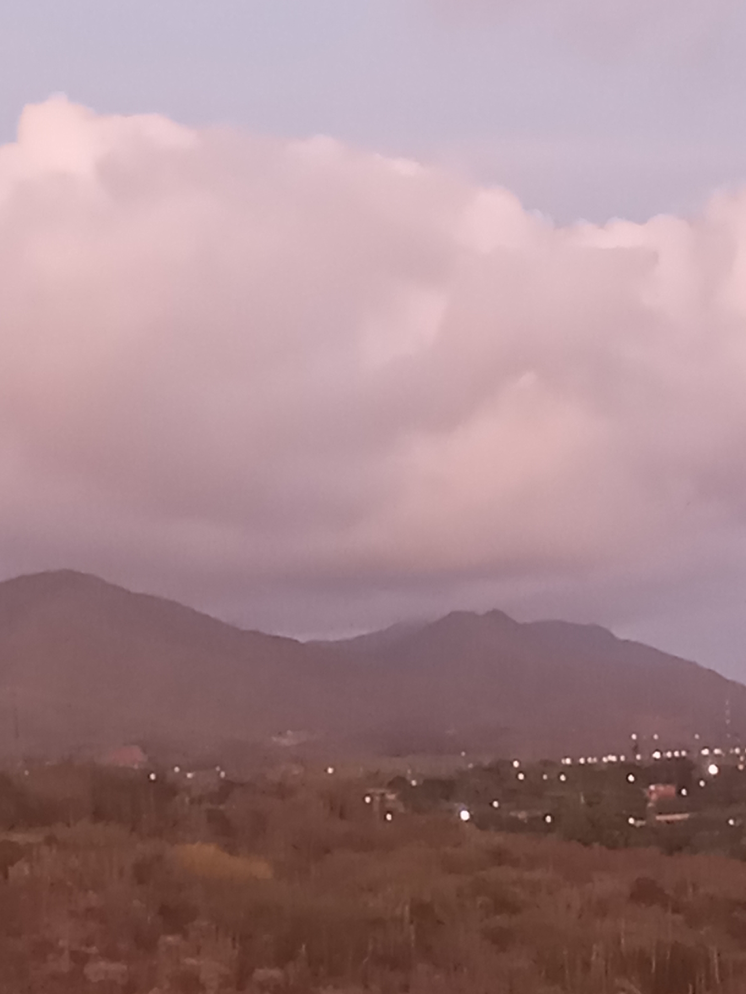 The image shows a landscape in Juan Griego, Venezuela, featuring mountains under a cloudy, pinkish sky. In the foreground, there is a field of low-lying vegetation. Scattered lights indicate the presence of a town or settlements in the middle ground. The mountains serve as a backdrop.