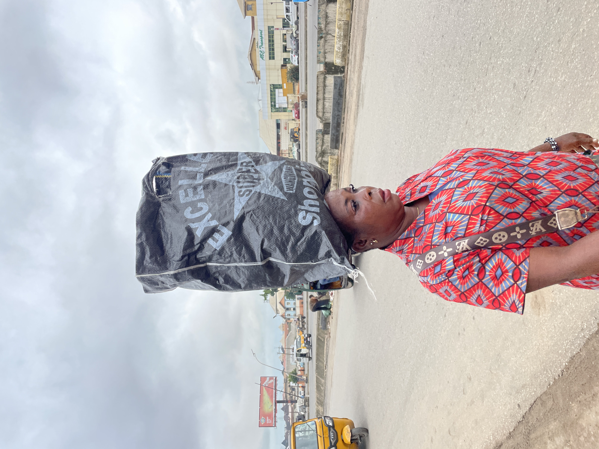 A medium shot captures a woman in Fegge, Nigeria, walking on a paved road. She is positioned in the foreground, with her body angled towards the right and her face turned to the left, looking ahead. The woman carries a large, black plastic bag balanced on her head. The bag is partially obscured by her head and is emblazoned with white lettering that reads "EXCELLENT SUPER WANGWANG Shopping." She is wearing a vibrantly colored, patterned shirt with red, blue, and white diamond shapes. A strap with a repeating pattern, possibly a designer logo, is visible across her shoulder.
The background depicts a busy urban street scene. A yellow auto-rickshaw is parked on the left side of the road. Further down the street, several other vehicles, including cars and a white van, are visible. The road is divided by a concrete barrier. In the distance, buildings with signage like "ABC Transport" can be seen. The sky is overcast with gray clouds. The overall environment suggests an active marketplace or transportation hub within the city.