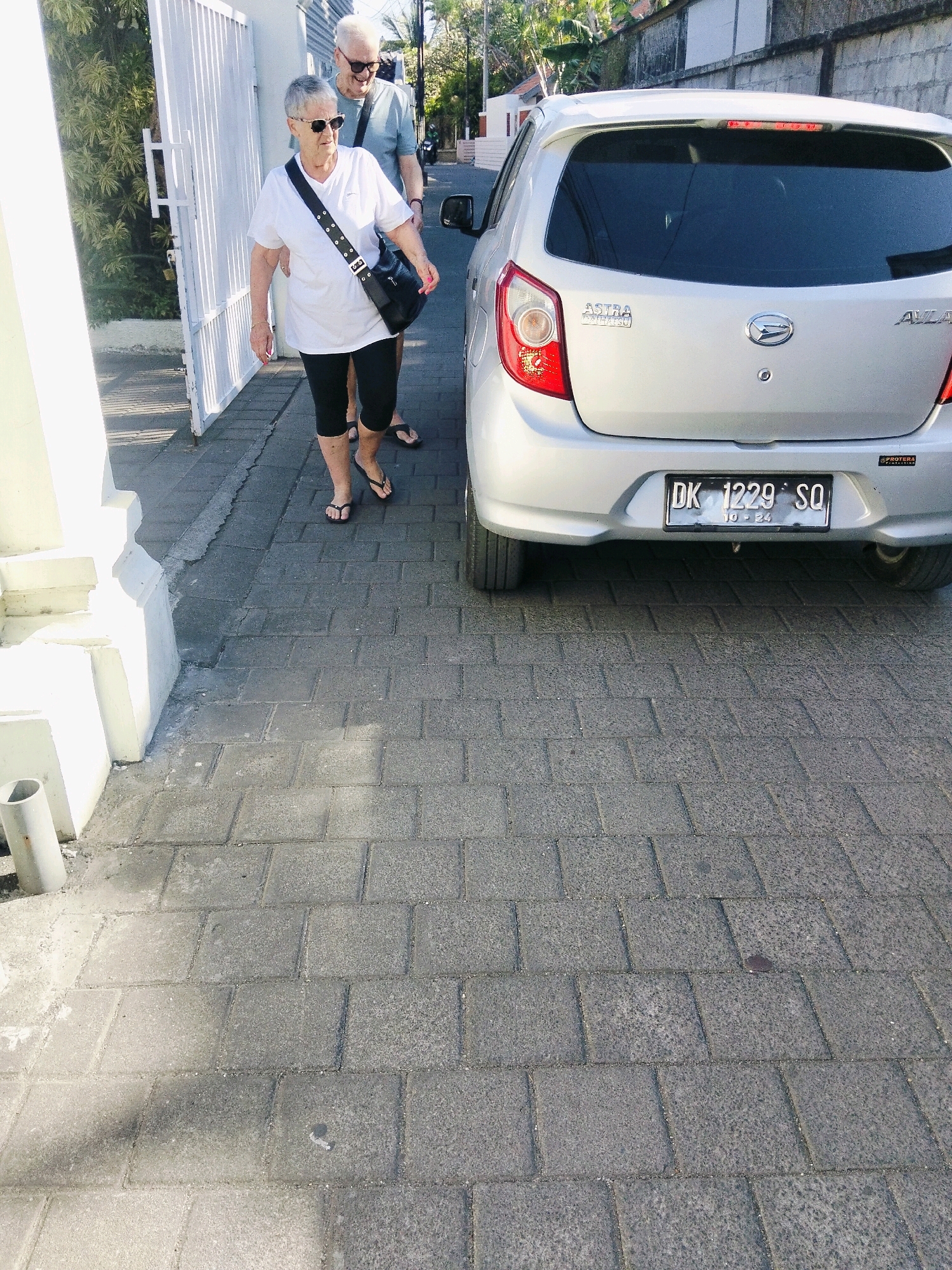 A senior Caucasian couple, identified by their light skin tone and hair color, walks on a brick-paved sidewalk in Kuta, Indonesia. The woman, on the left, wears a white t-shirt, black capri pants, and flip-flops. She carries a black crossbody bag. The man, on the right, is partially obscured by the woman. He wears a light blue t-shirt and dark shorts. Behind them, a silver Daihatsu Ayla car is parked. The car has a license plate that reads "DK 1229 SQ." The environment suggests a warm climate with sunlight casting shadows on the sidewalk. The buildings in the background are consistent with a tropical urban setting.