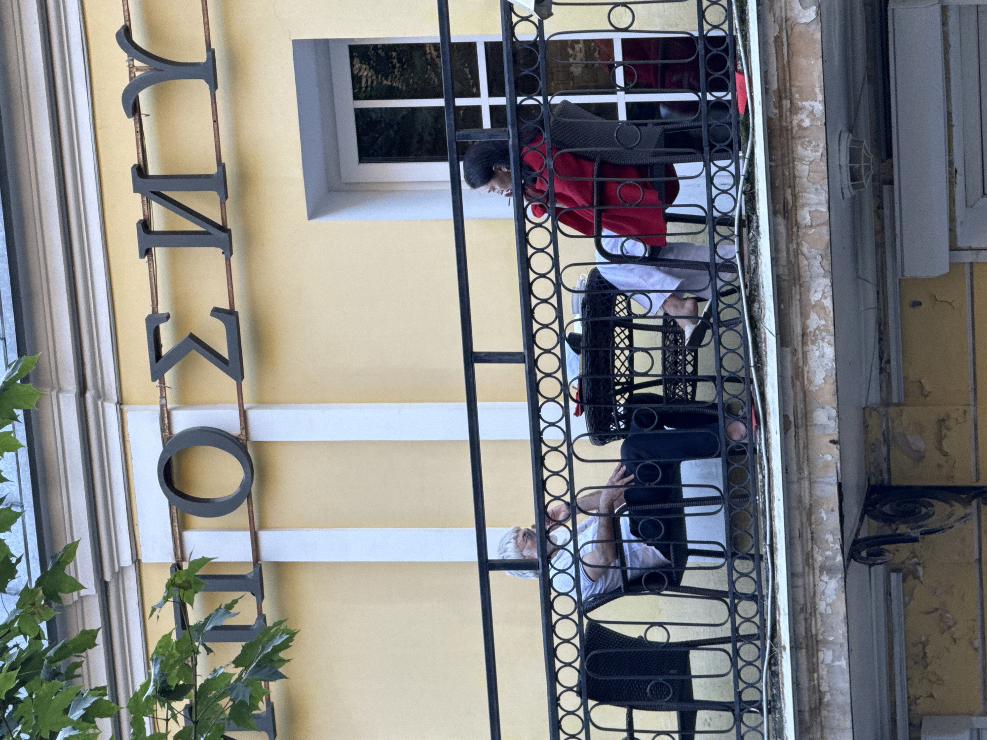 Two individuals are seated on a balcony of a building with yellow facade and ornate architectural details. Above them, the Cyrillic letters "ПОСУДУ" are mounted in dark metal. The left individual, appearing to be an older person with white hair, is seated in a dark chair, wearing a light-colored shirt and dark pants, and appears to be smoking. The right individual, a younger person with dark hair, is wearing a red jacket over white pants and is also seated in a dark chair, holding a cigarette. A small, dark table is placed between them, with items that may be cups or glasses on it. The balcony is enclosed by a black metal railing with a decorative circular pattern. In the background, a window with white mullions is visible. The image is taken from a slightly upward angle, with some foliage from a tree visible at the top left. The lower part of the image shows the peeling paint and weathered exterior of a lower level of the building, suggesting an older structure. The architectural style and lettering are characteristic of historic buildings in Saint Petersburg, Russia.