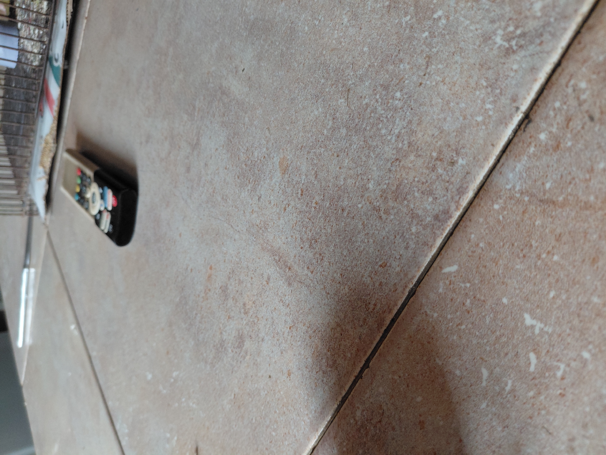 The image displays a close-up, angled view of a tiled surface, likely an indoor floor. The tiles are textured, appearing in shades of light brown and beige, with irregular patterns of darker speckles and lighter, whitish patches, resembling natural stone. A dark, distinct grout line runs diagonally through the middle of the frame, separating the tiles.
On the left side of the image, a remote control is partially visible, lying on its side on the tiled surface. It features a black body and a lighter-colored top, with an array of small, colorful buttons, including red, green, blue, yellow, and white. The buttons' labels are not discernible due to the slight blur and angle. Further to the extreme left, out of focus, parts of a metal cage or rack and a patterned bag or package are visible, suggesting the tiles are part of a domestic setting.
No people are present, and no particular activity is taking place. The scene is static, with objects resting on the surface. There are no notable time-of-day or weather cues, as the setting appears to be indoors. No visible text can be clearly read on any of the objects. The location context provided is Santa Ana, Venezuela.